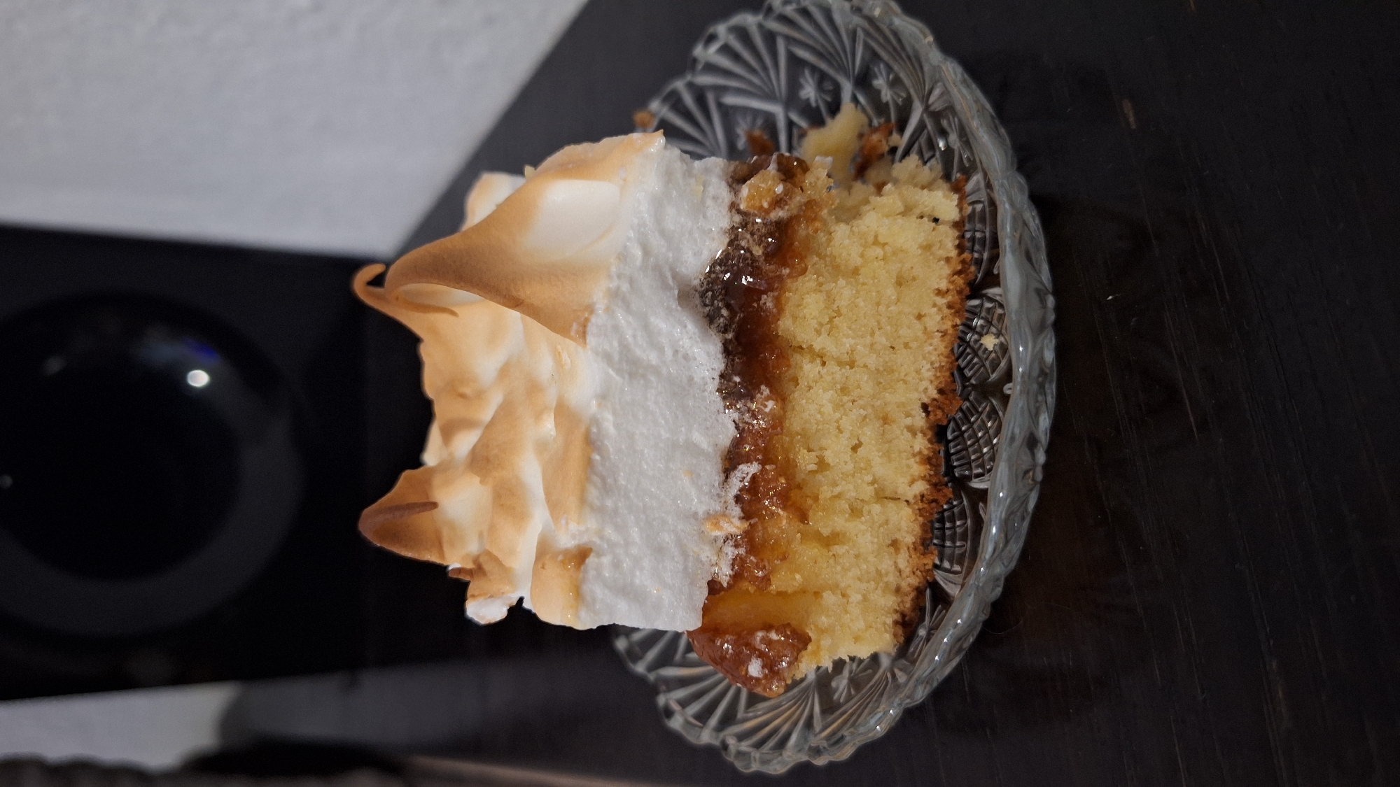 A slice of cake sits on a decorative glass plate on a dark, possibly wooden, surface. The cake has a layer of light-colored sponge, a jam-like filling, and a topping of browned meringue. The cake slice is placed to the right of the frame, with a dark reflective surface visible on the left. The overall lighting is subdued. It is not possible to determine the location of the image, including the city of Blagoevgrad, Bulgaria, from the visual content provided.
