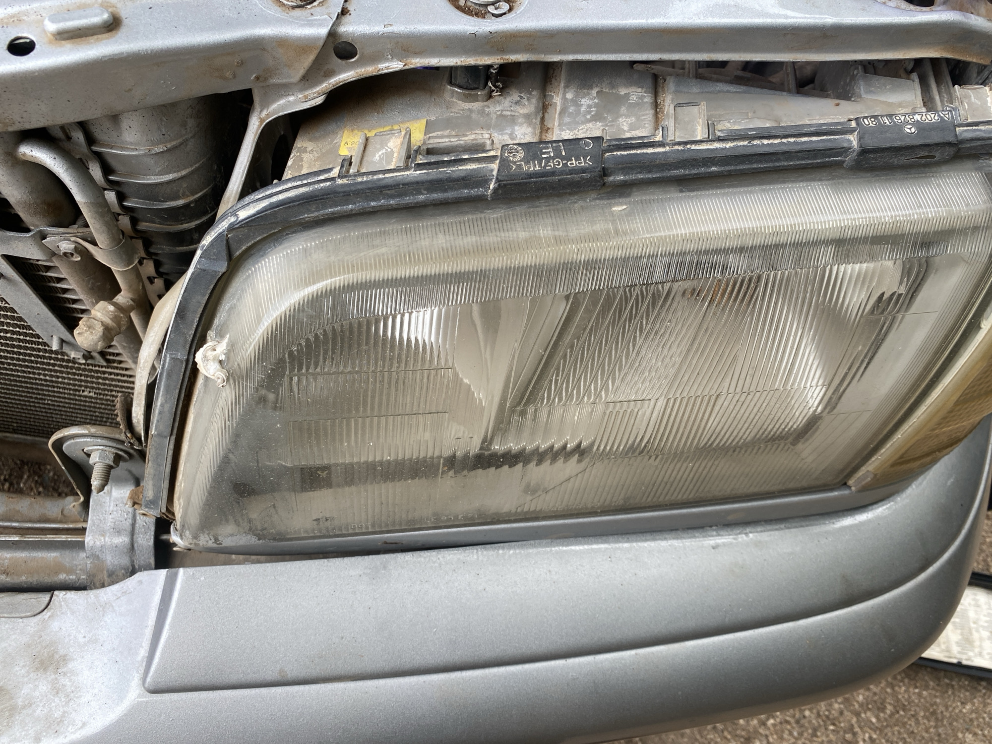 The image shows a close-up of a silver car's headlight assembly. The headlight lens appears somewhat cloudy and shows signs of wear. Attached to the headlight housing is a black plastic trim piece with text molded into it. Visible text includes "A 202 826 11 80", ">PP-GF/TPE<", and "OLE". Other markings include "A-20" on a yellow sticker.
Behind the headlight, parts of the car's engine bay are visible, including radiator fins, metal brackets, and tubing. The overall scene suggests a vehicle that has been exposed to the elements, with dust and grime present on the components. The lighting indicates it is daytime, but there are no specific cues about the weather or time of day. The context of Jalingo, Nigeria, suggests an outdoor setting, likely a repair shop or the car's usual parking location. No people are visible in the image.