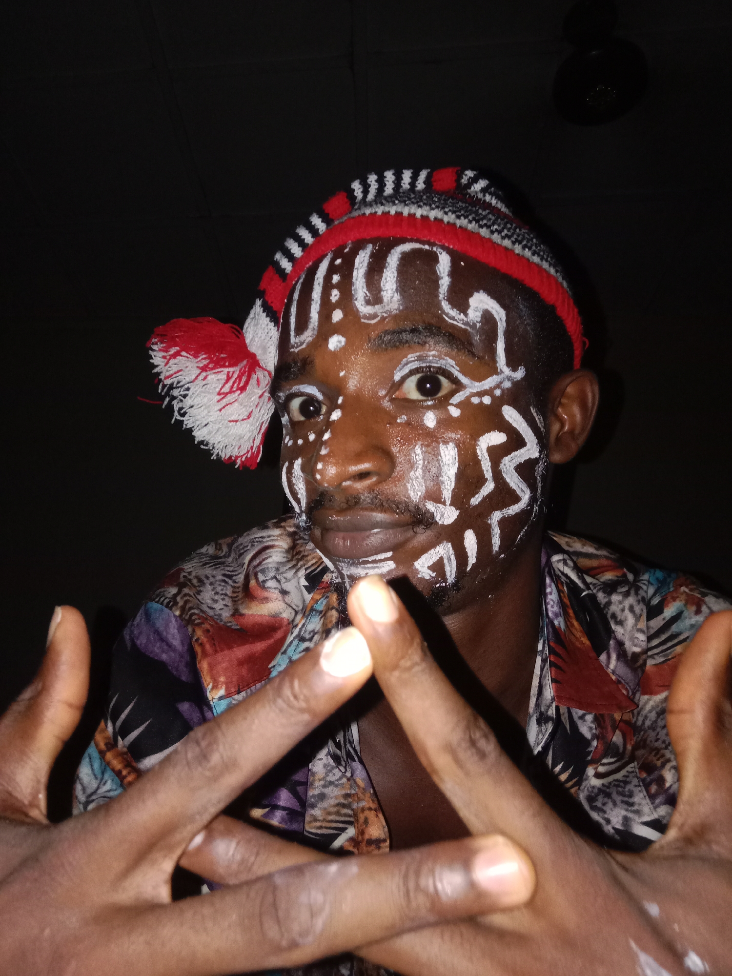 A medium close-up portrait captures a person with dark skin, their face elaborately decorated with white paint in tribal-like patterns of lines, dots, and swirls. Their eyes are wide open, directly engaging the camera, conveying an intense gaze. A subtle mustache and chin beard are visible.
The individual wears a distinctive knitted cap featuring red, white, and black stripes, adorned with a fluffy red and white pompom on the left side of their head. They are dressed in a brightly patterned shirt with a mix of colors including purple, blue, red, orange, black, and white, showcasing abstract or animal print designs.
Both hands are raised in front of the person, fingers spread apart to form a triangular or "X" shape, with some white markings visible on the right hand. The fingertips appear lighter in color. The background is very dark, suggesting a low-light indoor setting or nighttime. A faint circular object, possibly a light fixture or ceiling fan, can be vaguely discerned in the upper right. The image portrays a cultural performance, artistic expression, or ritual. No visible text is present.