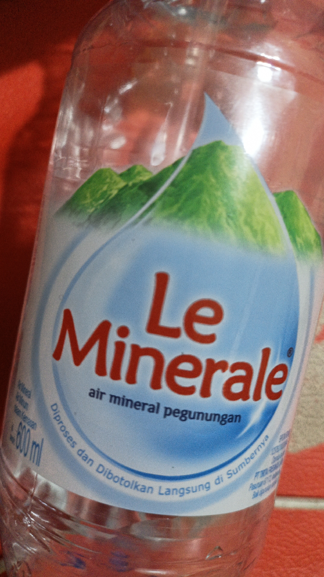 The image shows a close-up of a plastic bottle of "Le Minerale" bottled water. The label is primarily blue and white with red text. The prominent text on the label reads "Le Minerale" in large red letters. Below this, in smaller blue text, it says "air mineral pegunungan," which translates to "mountain mineral water." There are green graphics of mountains on the upper part of the label. The bottle appears to be partially filled with water. The background is a blurred red surface, possibly a tablecloth or a wall. There is no indication of the city of Palohlada, Indonesia, in the image itself. The scene is a still life product shot of a water bottle.
