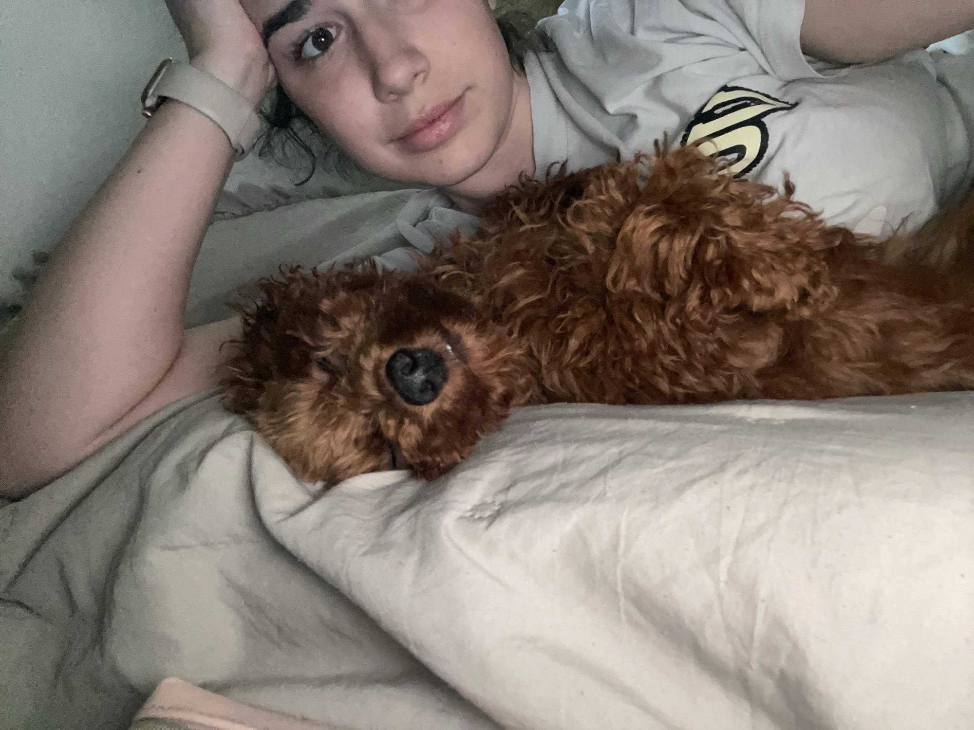 A woman and a brown dog are lying on a bed. The woman is on her left side with her head propped up by her left arm. She is wearing a light-colored t-shirt and a watch. The dog is lying next to her, facing the camera. The bed has light-colored sheets. The location is not discernible from the image.
