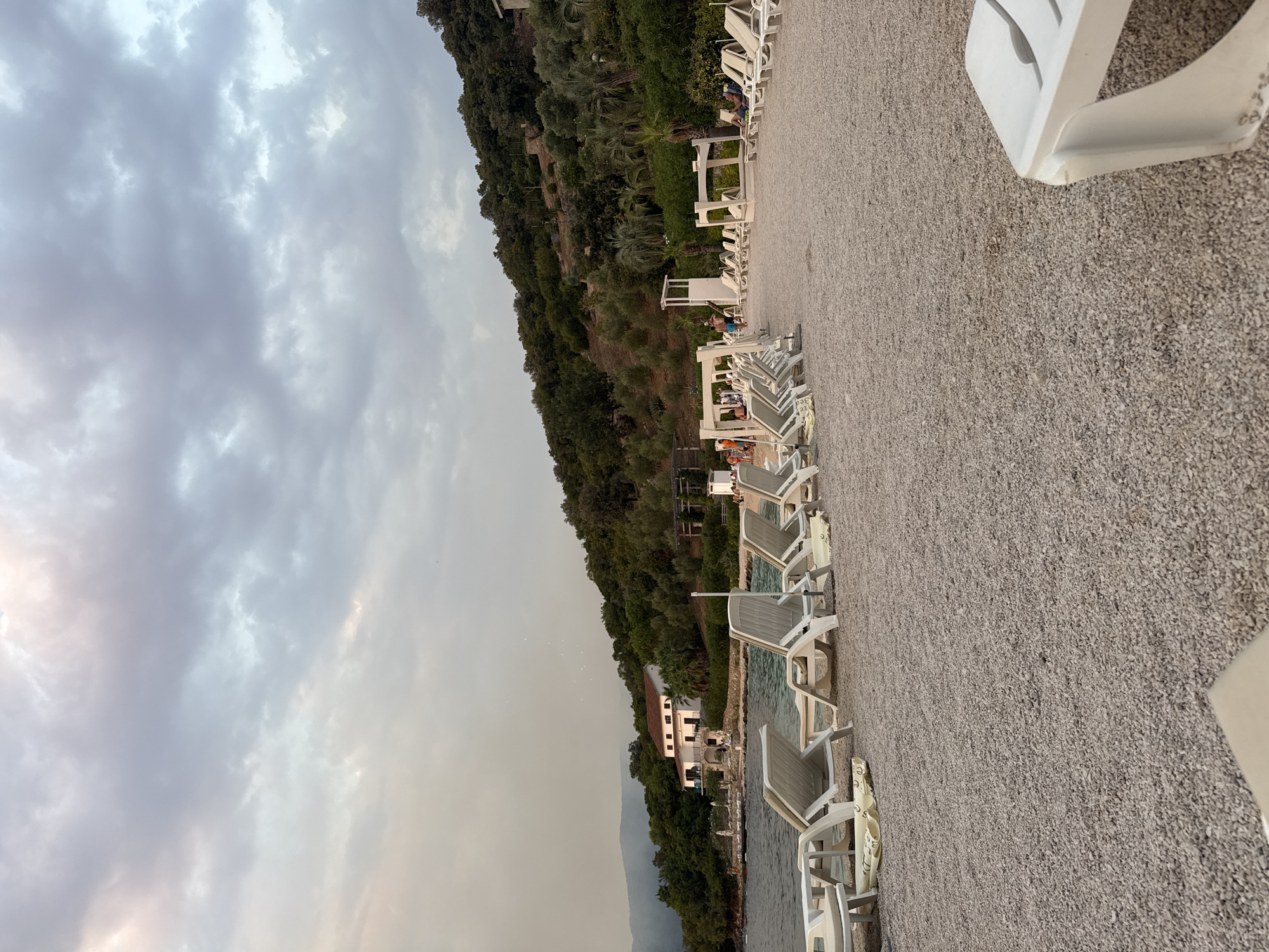 The image depicts a wide-angle view of a pebble beach with rows of empty white lounge chairs. In the foreground, a white plastic lounge chair is partially visible on the right. The beach slopes gently towards the water, which is calm and reflects the overcast sky. A white building with a red-tiled roof is situated on the left side of the image, nestled among lush green trees and vegetation that cover a hillside. Further up the hill, there are more trees and some stone structures. Several small white canopies or gazebos with white supports are set up on the beach. In the middle ground, a few people are visible walking and standing near the canopies. The sky is filled with dramatic, heavy gray clouds, with hints of pale light breaking through in some areas. The overall impression is a serene, slightly moody beach scene.