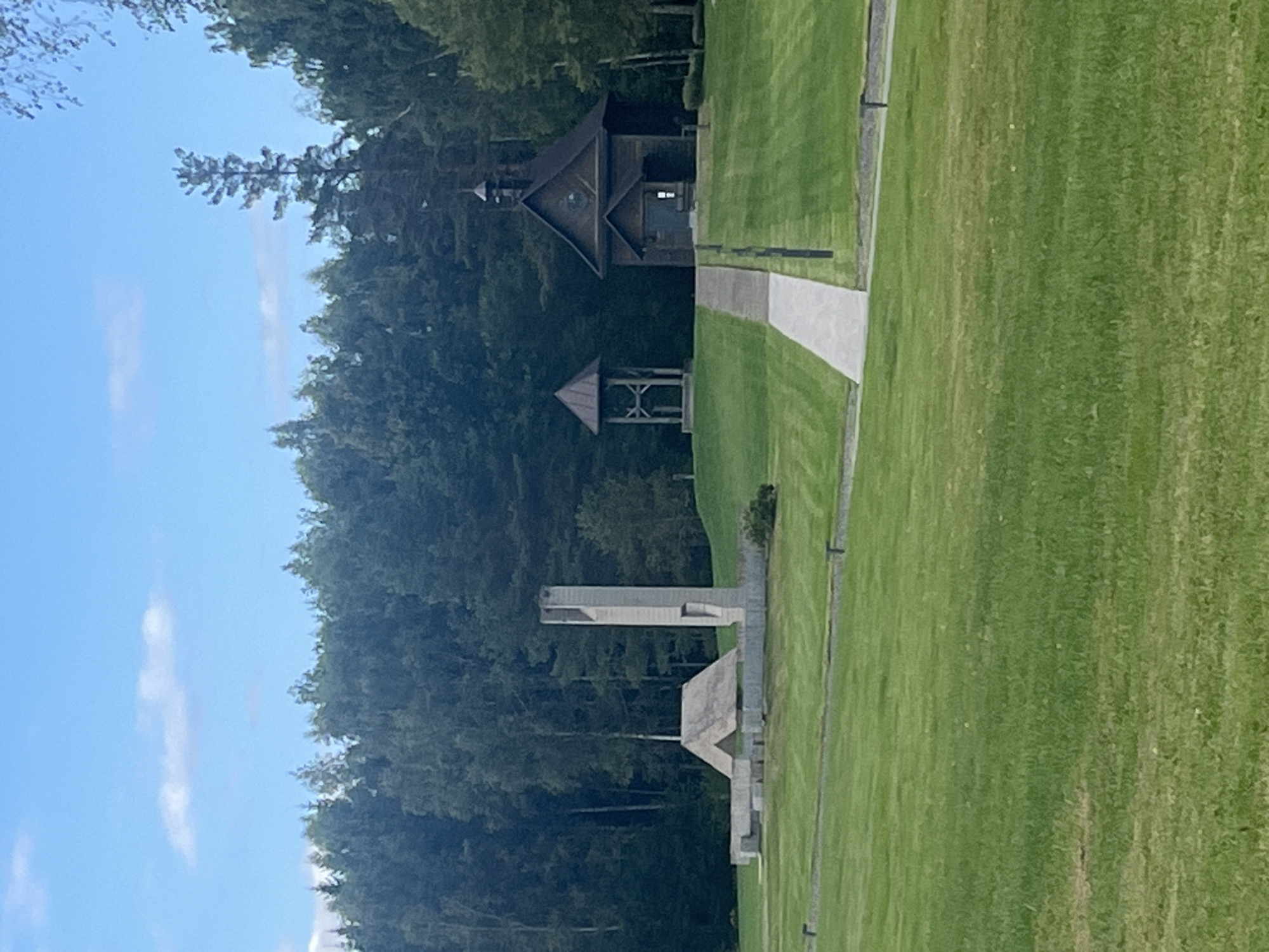 The image depicts a serene outdoor memorial site situated amidst a lush green lawn and a dense forest backdrop under a partly cloudy sky. In the foreground, a neatly manicured lawn is divided by a paved walkway leading towards two distinct structures on a slightly elevated grassy area. To the left, a monumental structure composed of concrete elements, including a tall, slender tower with an opening and a lower, angled roofed section, stands as a focal point. To the right, a charming wooden chapel with a steep pitched roof and a small bell tower is visible, alongside a separate, open-sided bell tower. The overall impression is one of solemnity and remembrance, with the natural surroundings contributing to a peaceful atmosphere. The location appears to be the Khatyn Memorial Complex, a significant historical site in the village of Khatyn, within the Kamenetsky District of Belarus. There are no people visible in the image.