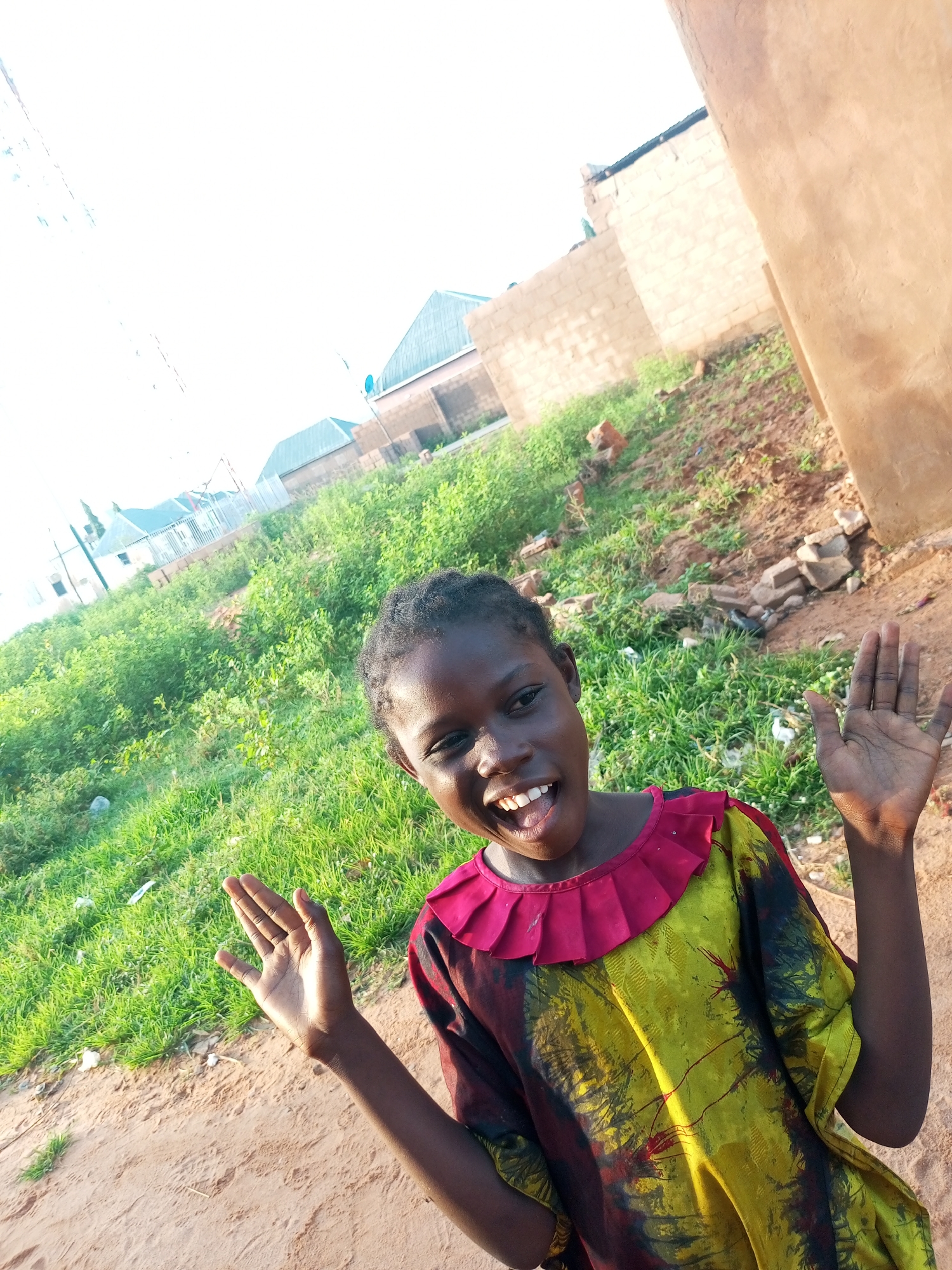 An African child, appearing to be a girl, smiles widely with her mouth open, showing her teeth, and raises her hands in greeting. She is wearing a loose-fitting, colorful garment with a ruffled red neckline and a pattern of yellow, black, and red. The child is standing on a sandy dirt path. Behind her, there is an area of lush green vegetation and some scattered bricks and debris. In the background, several buildings with brick walls and sloped roofs are visible. The sky is bright and overcast, suggesting it might be daytime. The scene is consistent with an outdoor environment in a developing urban or rural area, possibly in Tempure, Nigeria.