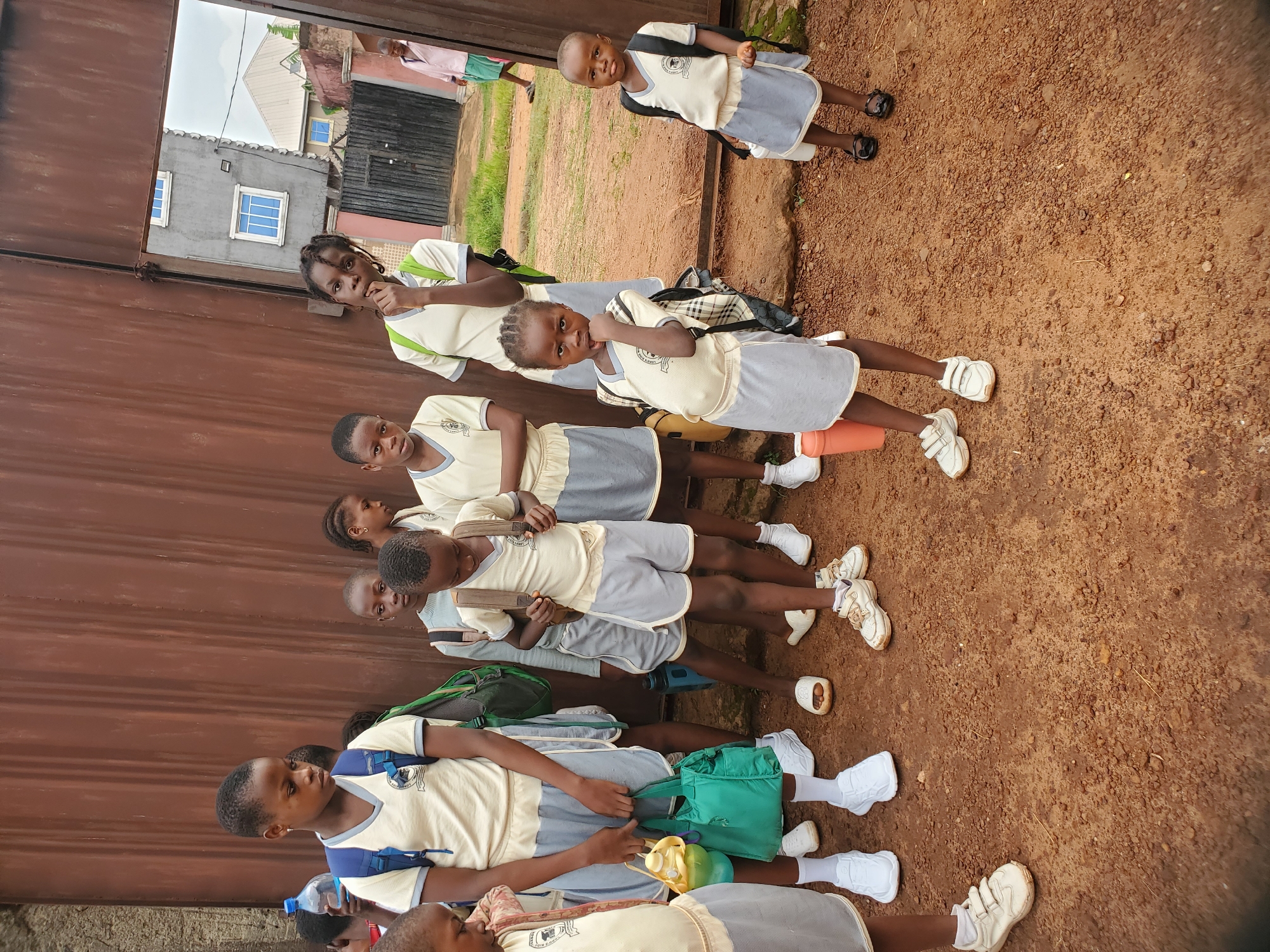 A group of approximately ten children, all wearing similar school uniforms (light yellow shirts and gray shorts or skirts), stand or walk alongside a corrugated metal fence. The setting appears to be in Ukwu-Abwa, Nigeria, based on geographical reference. The children are positioned on a dirt or soil surface, with some carrying backpacks. A few buildings and foliage are visible in the background. Some of the children are looking directly at the camera. One is standing apart from the others on a slight embankment.