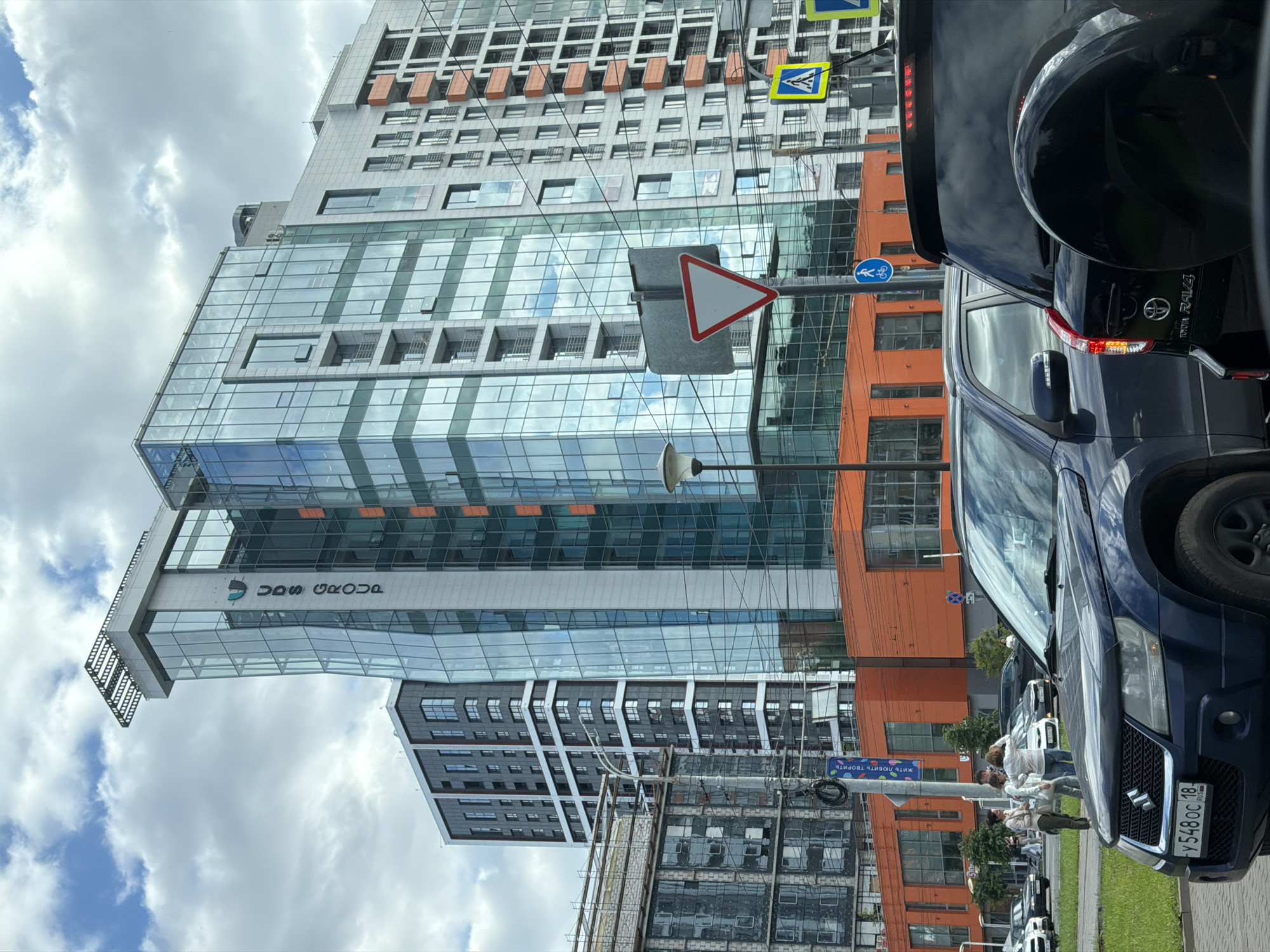 This image captures a street view in Izhevsk, Russia, focusing on modern architecture and urban traffic. In the foreground, a dark blue Suzuki Grand Vitara SUV is parked, facing towards the viewer, with its license plate clearly visible: 'y 548 oc 18 RUS'. To its right, the rear of a black Toyota RAV4 is visible.
Behind the vehicles, a striking modern office building with a glass facade dominates the scene. The building features sections with vibrant orange lower levels and upper floors clad in reflective blue-tinted glass panels. The text "UDS GROUP" is prominently displayed vertically on the left side of the glass facade. Adjacent to this building are other high-rise structures, including a darker-colored residential building with numerous windows and balconies, and another partially constructed building with scaffolding.
The street is lined with trees, and a traffic light pole with various road signs is present. One prominent sign is a red-bordered yield sign, and another is a yellow diamond-shaped sign with a pedestrian and bicycle symbol. The sky is partly cloudy, indicating a temperate day. Several people are seen walking on the sidewalk and near the buildings, some appearing to be pedestrians waiting or conversing. The overall impression is of a contemporary urban environment with active traffic and new construction.