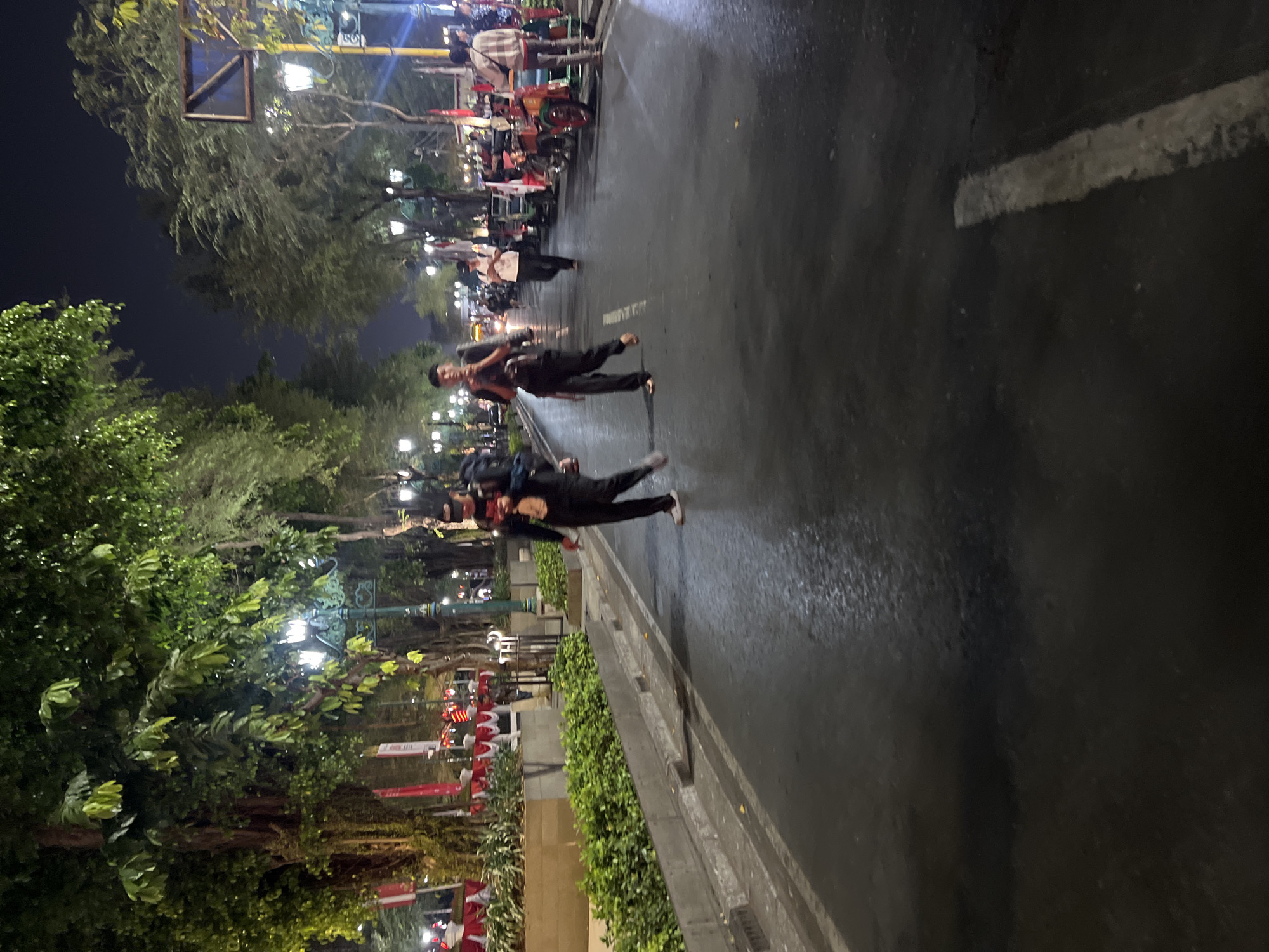The image shows a street scene at night in the city of Yogyakarta, Indonesia. The street is wet and reflects the ambient light from lampposts and vehicles. On the left side of the street, there are trees with lush foliage, and a row of red and white banners. Two individuals are walking on the street, one in the foreground on the right and another slightly further back on the left. The person in the foreground is wearing dark clothing and a baseball cap, and appears to be looking at their phone. The person on the left is also wearing dark clothing and carrying a bag over their shoulder. Several motorbikes and what appear to be pedicabs are parked along the right side of the street, some adorned with flags. The overall atmosphere is that of a busy street, possibly a marketplace or a promenade, illuminated by artificial lights against the dark night sky.