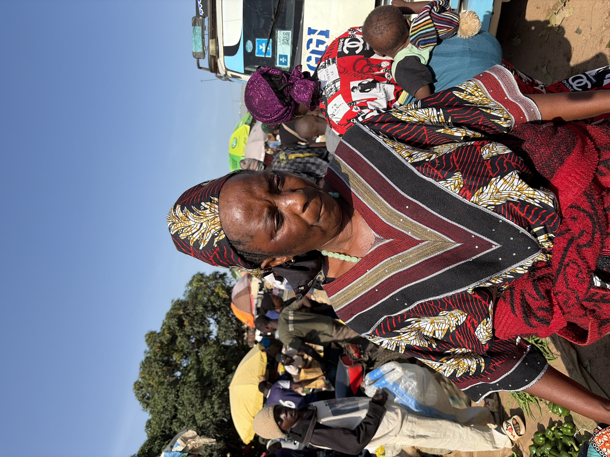 A close-up, waist-up shot shows a Black woman wearing a colorful, patterned headscarf and dress. The dress has a V-neck with striped patterns in black, red, white, and gold, accented with small silver beads. A green beaded necklace is visible around her neck. She is in an outdoor marketplace in Gura Top, Nigeria, with a clear blue sky above.
To her right, another Black woman wearing a purple headscarf and a red shirt with white graphics is seen from the back, carrying a baby on her back. The baby is wearing a striped hat and blanket. Behind them, a white vehicle with "LAGOS" written on its license plate is partially visible, along with other market stalls, people, and trees. To the left of the main subject, a Black person in a beige outfit and a light brown beanie is visible, carrying a white plastic bag. A pile of green bell peppers is on the ground in the foreground. The ground appears to be dirt.