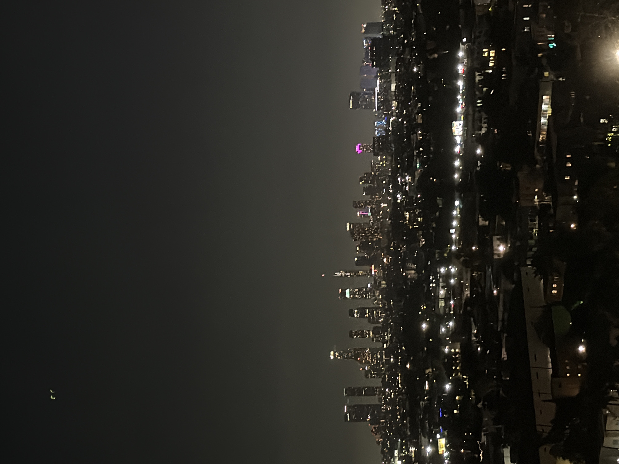 A nighttime aerial view of the Los Angeles skyline is captured, with the downtown area illuminated by countless lights. The buildings vary in height and architectural style, with some displaying colorful illuminated tops, such as purple and pink lights on a few of the towers. In the foreground, the lights of residential areas and lower-rise structures extend towards the viewer, creating a dense urban landscape. Two small, bright, crescent-shaped lights are visible in the dark sky in the upper left quadrant of the image, their nature undetermined. The overall atmosphere is one of a sprawling, vibrant city at night.