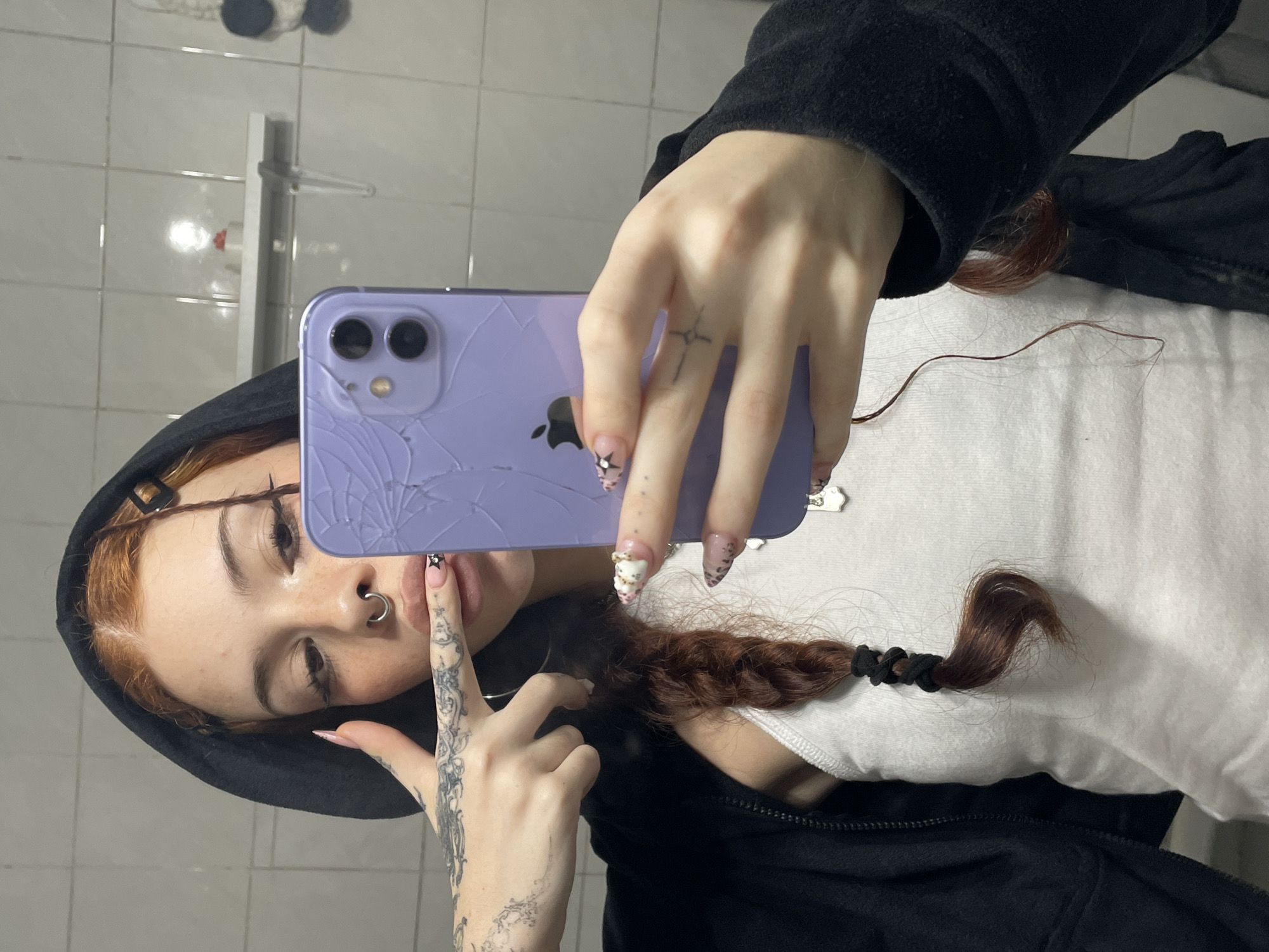 A young person is taking a selfie with a lavender iPhone that has a cracked screen. The person has reddish-brown hair, some of which is braided, a nose ring, tattoos on their hands and arms, and is wearing a black hooded jacket over a white tank top. The background shows a tiled wall, suggesting a bathroom. There is no indication of Buenos Aires, Argentina.
