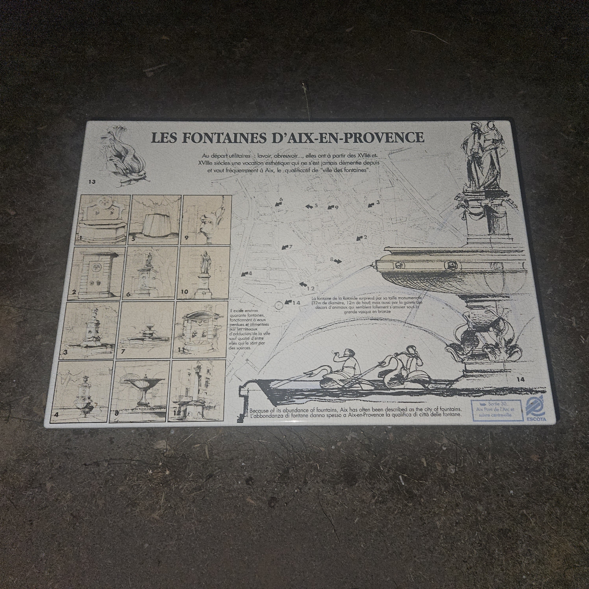 The image shows a plaque detailing fountains in Aix-en-Provence, France. The plaque features drawings and text in French, English, and Italian. It includes a map of the city with numbered locations of fountains and individual illustrations of various fountains. A large drawing at the bottom depicts people on boats interacting with a fountain. The plaque is positioned on a dark, rough surface, likely the ground, with some debris and dry plant matter visible. The lighting is subdued, casting shadows on the scene. There are no people visible interacting with the plaque or in the immediate vicinity. The plaque itself appears to be made of a hard material, possibly ceramic or porcelain, with a slightly reflective surface. There is no indication of the specific location within the city of Rousset, France, as the plaque is about Aix-en-Provence.