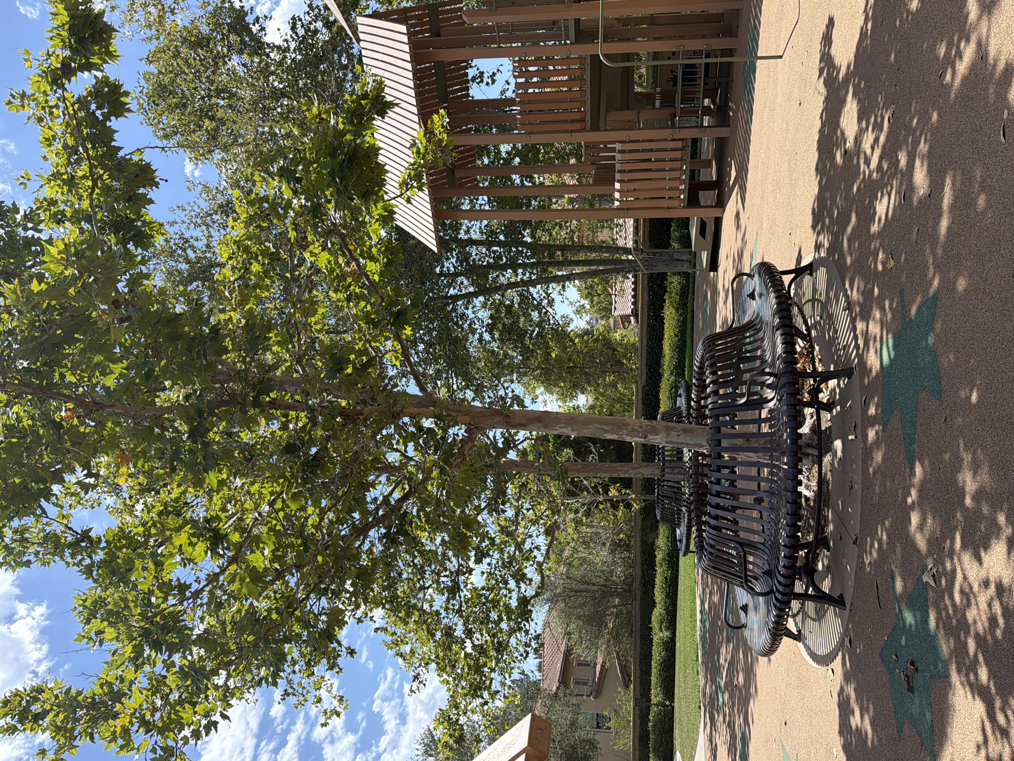 The image captures a sun-drenched park scene, likely located in the city of Irvine, United States of America, on a clear day with scattered clouds. In the foreground, two circular, dark metal benches encircle the base of a large sycamore tree. The benches have a slatted design and appear to be part of a playground or recreational area. The ground is a light brown, textured surface with faint, green star-shaped patterns embedded within it, casting dappled shadows from the tree's leaves.
To the right, a multi-level wooden playground structure stands prominently. It features a slatted roof, railings, and platforms, suggesting it is designed for children's play. Behind the playground and benches, a dense line of green trees and shrubs creates a natural backdrop. In the distance, beyond the foliage, residential buildings with terracotta tile roofs are visible, indicating the park is situated within a developed neighborhood. The overall atmosphere is tranquil and inviting, bathed in natural light. There are no people or animals visible in the scene.