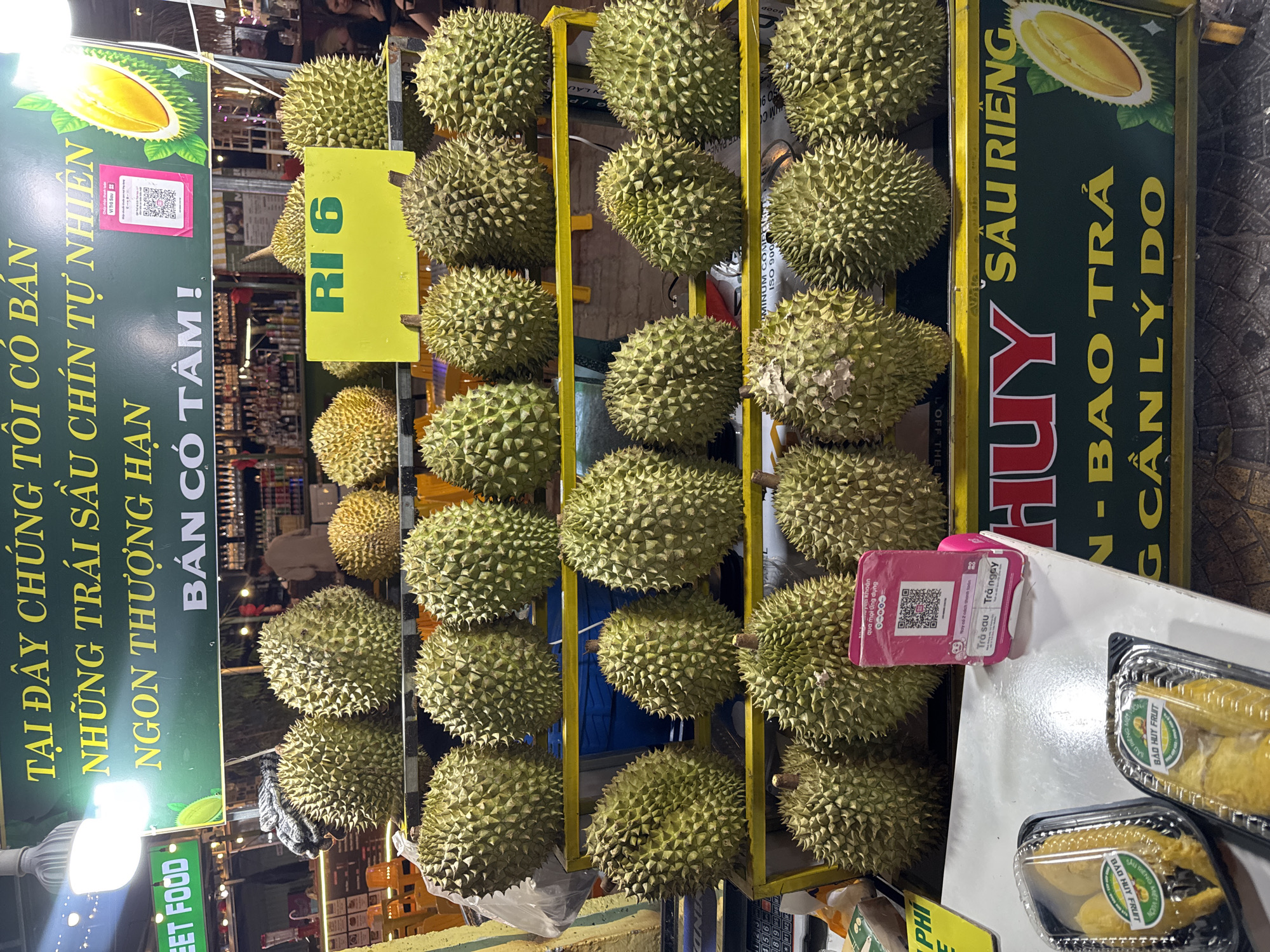 Here's a detailed description of the image:
**Scene:** An outdoor street food market stall specializing in durian, the thorny tropical fruit known for its pungent smell. The scene appears to be in Vietnam, based on the language on the signs.
**Subjects:** Stacks of durian fruit, arranged on a yellow metal grid. Signs promoting the durian, including prices. Packaged durian fruit is available for sale as well.
**Objects:**
* Many whole durians displayed.
* Signs in Vietnamese advertising the durian, stating "Only sell naturally ripened durian," "Delicious, premium" and "Selling with heart!"
* Price tag: "Ri 6" on yellow tag.
* Pink QR code scanner.
* Containers of pre-packaged, cut durian.
* Lights and other market stalls visible in the background, suggesting a busy street food area.
**People:** Not clearly visible. There is an obstructed view of a person in the background by food.
**Environment:** An outdoor market setting, likely at night, given the artificial lighting.