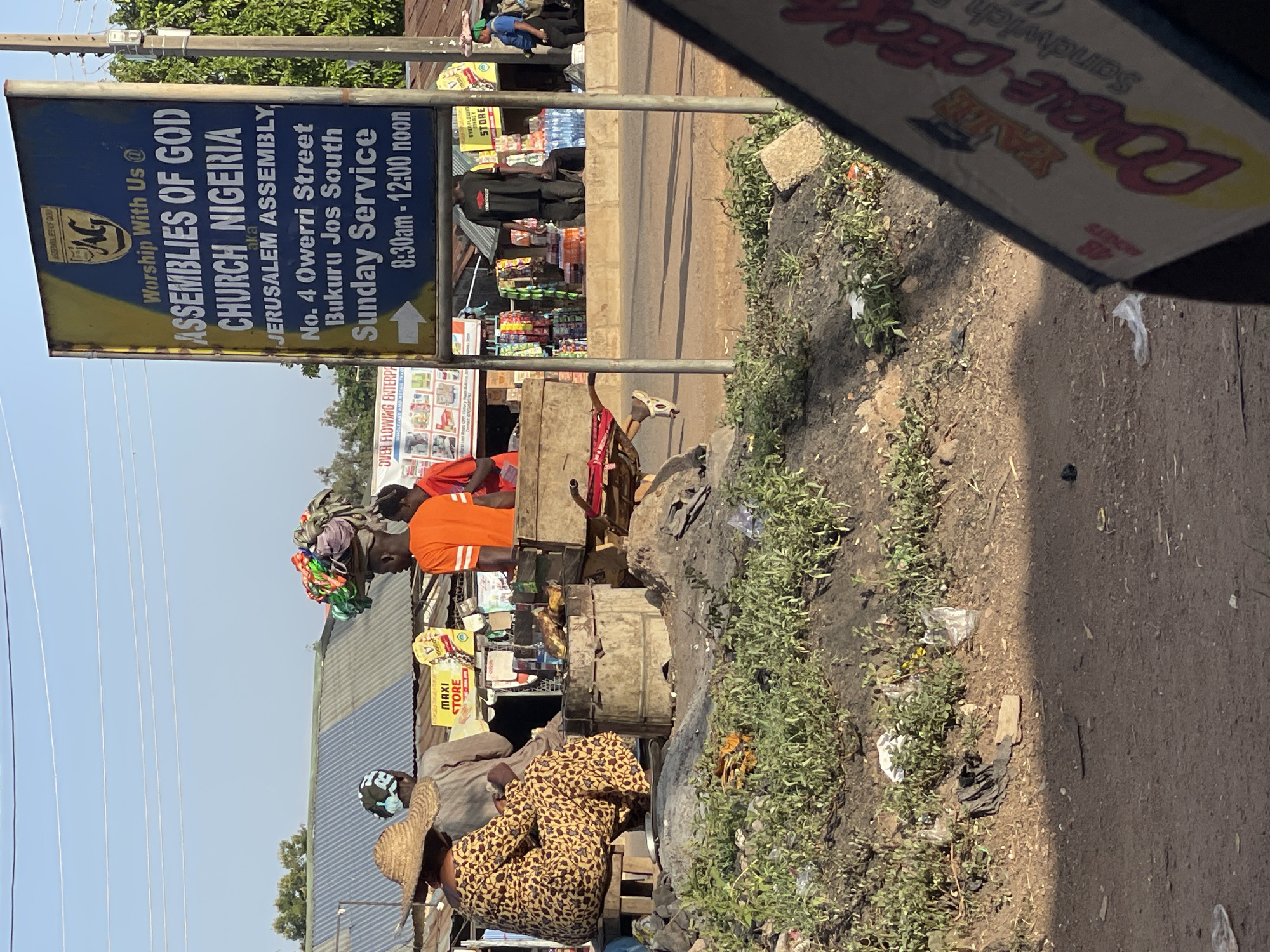 A vertical, outdoor daytime shot shows a street scene in Bukuru, Nigeria with a church sign, market stalls, and passersby. In the foreground, a sign partially obscures the view, displaying "DOUBLE-DECK Sandwich." Adjacent to it, roadside vegetation and litter clutter the edge. The church sign prominently displays "ASSEMBLIES OF GOD CHURCH NIGERIA, Jerusalem Assembly, No. 4 Owerri Street, Bukuru Jos South, Sunday Service 8:30am-12:00 noon." Further down the street, market stalls are visible with vendors and customers. One man wears an orange shirt, and another woman wears a leopard-print dress and a straw hat. Goods are displayed at the market, and people are engaged in commerce.