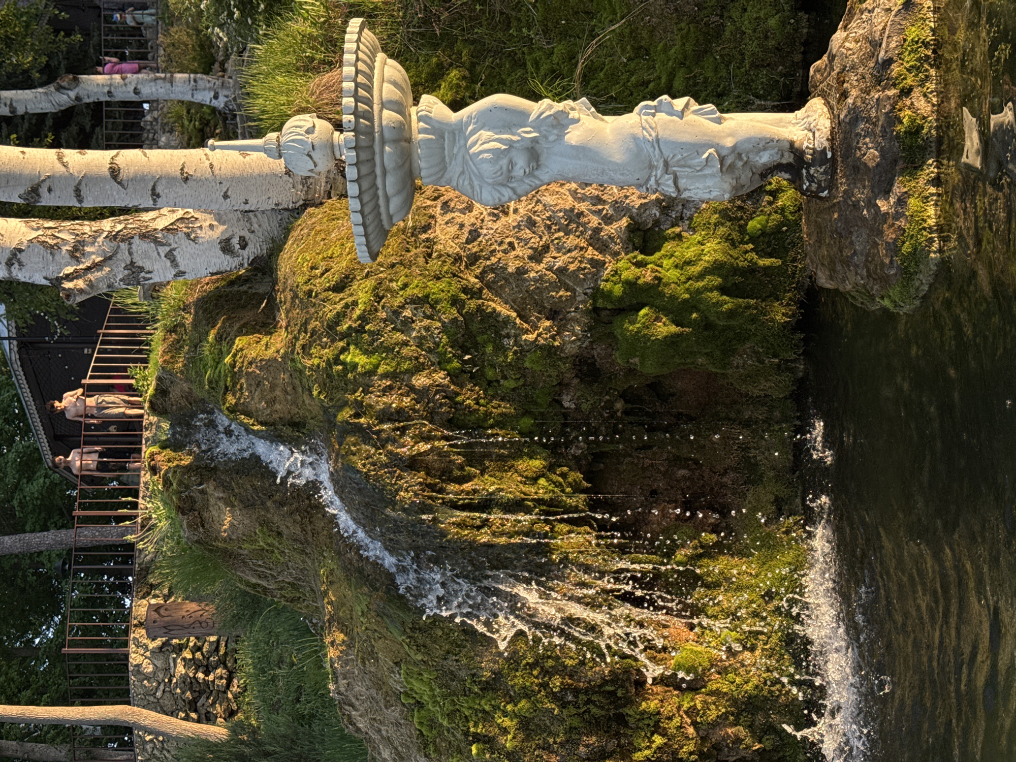The image captures a scene in Krasny Klyuch, Russia. A moss-covered rocky formation with a small waterfall is prominent. A classical-style sculpture rests atop the rock. Water flows from the sculpture into a dark pool. A man reclines on a platform supported by birch trees and a metal railing is visible in the background. A wooden staircase is near the platform.