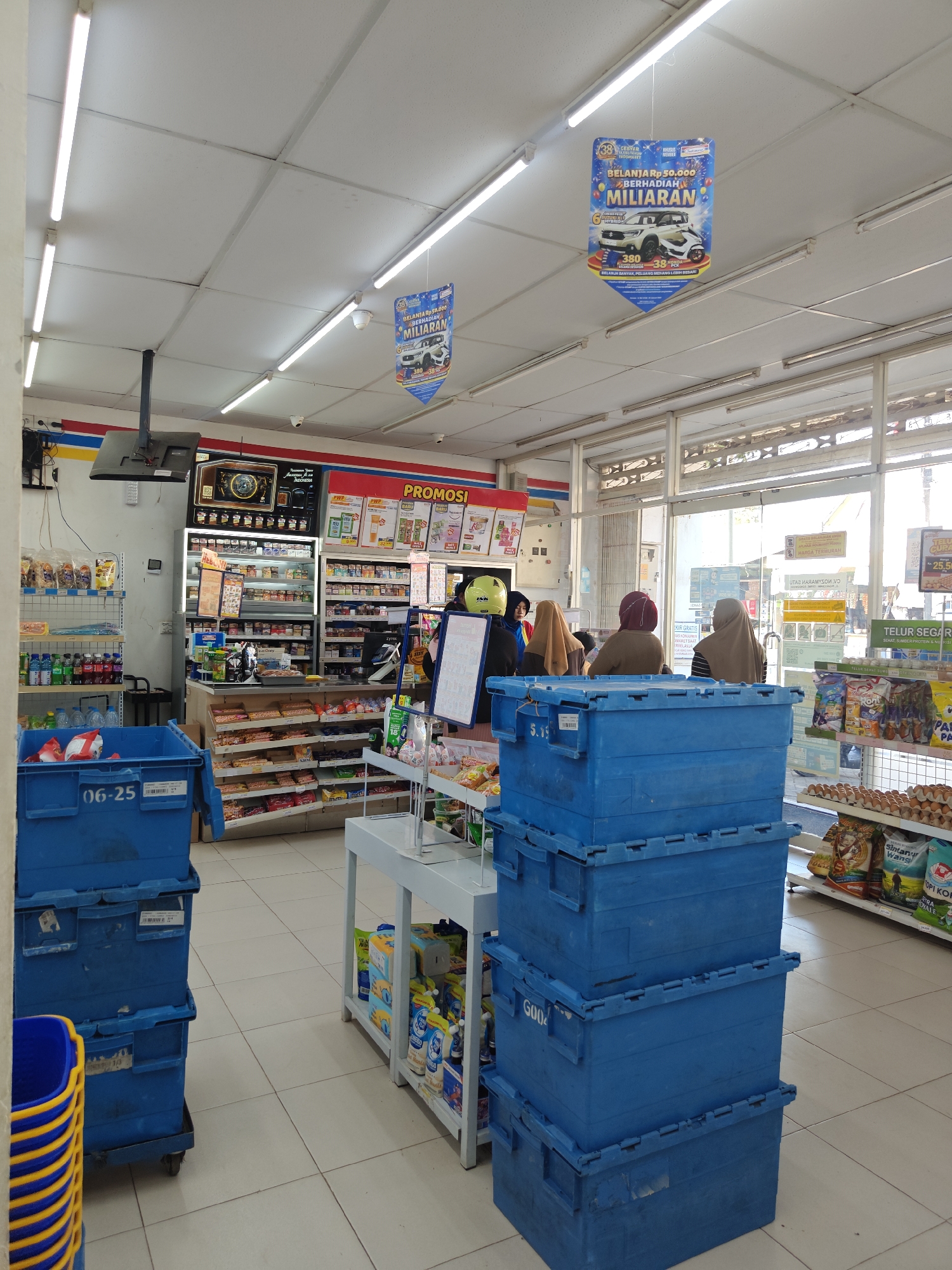 The image shows the interior of a convenience store, possibly Indomaret, located in Klampokan, Indonesia. The store is brightly lit by fluorescent ceiling lights and displays a variety of products on shelves. In the foreground, to the left, are stacked blue plastic crates, one with the label "06-25". Further into the store, a checkout counter is visible with several customers queuing. Three women are standing at the counter, two of whom are wearing headscarves. Behind the counter, shelves are stocked with packaged goods. A promotional banner hangs from the ceiling, advertising a shopping promotion with prizes. To the right, a shelf displays a variety of eggs and other grocery items. The floor is tiled. The store appears to be clean and organized.