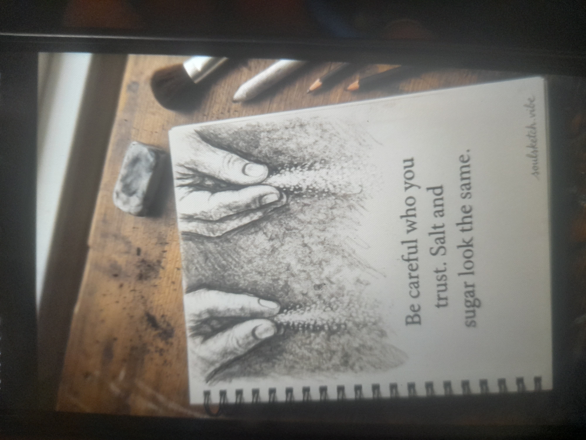 The image shows a close-up shot of a drawing on a notepad with the quote "Be careful who you trust. Salt and sugar look the same." The drawing depicts two hands holding a line of what appears to be salt or sugar. The notepad is on a wooden surface, and there are drawing supplies such as brushes, pencils, and an eraser nearby. The location of the image is not Akwanga, Nigeria, as the scene provides no discernible link to that location.