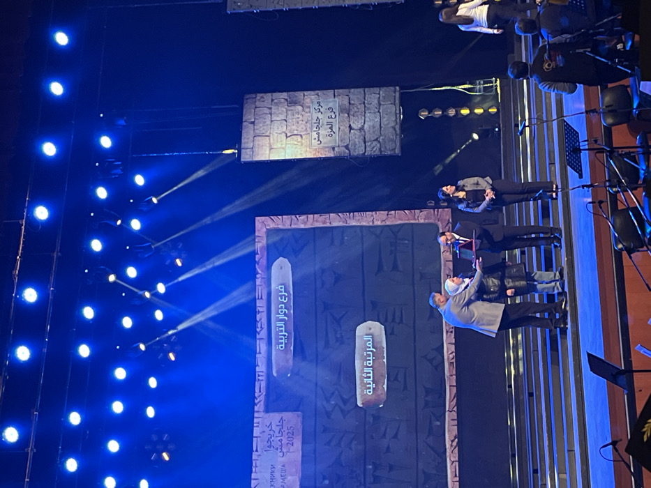 The scene depicts an awards or recognition ceremony taking place on a brightly lit stage within the city of Al-Qanawat Municipality, Syria. The stage features multi-tiered steps and a large backdrop resembling ancient stone tablets or cuneiform, displaying Arabic text. The text on the left reads "خريجوا جلجامش 2025" (Graduates of Gilgamesh 2025), with "فرع دوار التربية" (Education Roundabout Branch) and "المرتبة الثانية" (Second Place) displayed centrally. On the right, a vertical banner reads "مركز جلجامش فرع المزة" (Gilgamesh Center Al-Mezzeh Branch).
In the center of the stage, four individuals are engaged in an interaction. A man in a grey suit jacket stands on the left, extending his right hand to shake the hand of a woman in a white hijab, black jacket, and blue jeans, who stands facing the audience's left. To her right, a man in a dark suit and glasses holds a red-cased object, possibly an award or plaque, presenting it. A woman in a black blazer and dark pants stands observing to the right of the presenter.
The stage is illuminated by numerous overhead blue lights and spotlights. In the foreground, an orchestra pit or performance area is visible, with multiple microphones on stands, hinting at musical accompaniment. Individuals are partially visible in this area, including one person wearing a dark jacket with a yellow emblem. On the far right of the stage, another woman in a white shirt and dark pants is descending the steps.