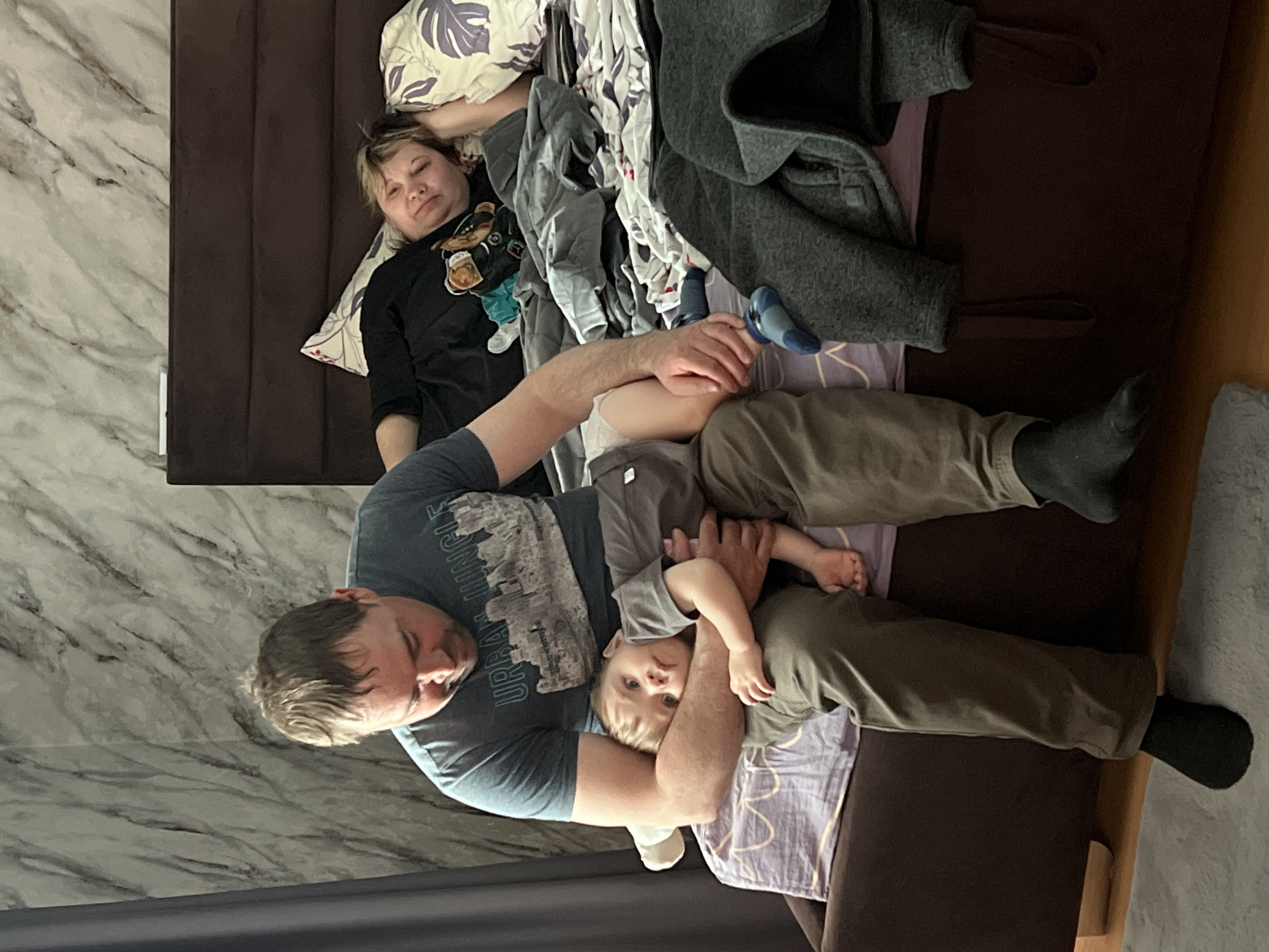 A photo taken indoors shows an adult man, an adult woman, and a young child lying together on a bed. The man is wearing a grey t-shirt and olive drab pants and socks. The woman is wearing a dark shirt with a bear design. The child is wearing a light grey shirt. The woman is asleep. The man is holding the child, and the child is looking at the camera. The bed is covered with a brown headboard and lavender sheets. Other items include a gray coat, clothes, and pillows. The backdrop includes a wall with a marble pattern. No visual elements suggest the location is Perm, Russia.