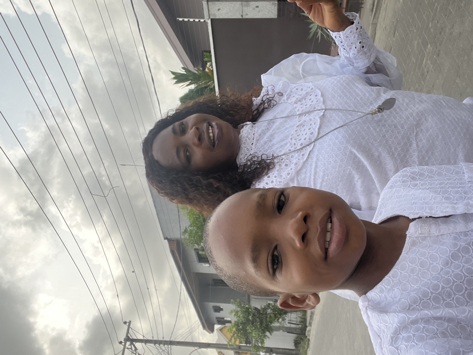 A photograph taken in Iranla, Nigeria, depicts an adult female and a child posing outdoors on a paved surface under an overcast sky. In the foreground, slightly to the left, a child with light brown skin wears a white, short-sleeved top with a circular textured pattern, looking directly at the camera with a slight smile. Behind and to the right, an adult female with dark, curly hair and light brown skin is visible from the chest up, smiling with her mouth slightly open, looking slightly off-camera to the right. She wears a white high-necked blouse featuring intricate eyelet lace details and ruffles on the collar and cuffs. A long, silver-colored necklace with an oblong, decorative pendant hangs around her neck. Her left hand, with fingers slightly splayed, is partially visible on the right side of the frame. The background features residential structures: a light-colored building with dark windows and a peaked roof is visible on the left, while a darker wall or building section topped with a security fence (electric fence insulators/wires) is present on the right. Green foliage is interspersed between the subjects and background buildings. A utility pole with multiple overhead wires is visible on the far left. The ground is a paved surface, consistent with a road or sidewalk.