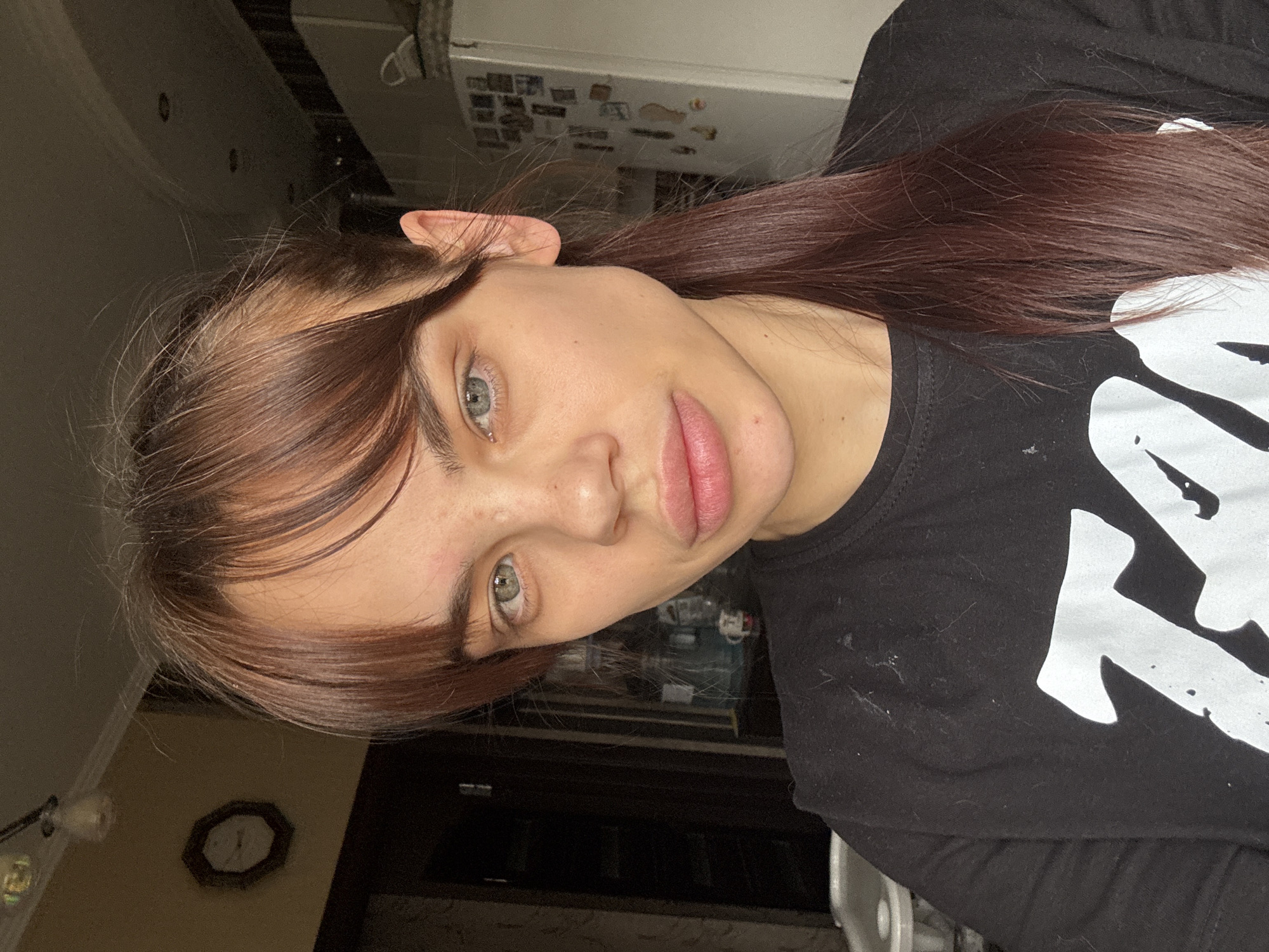 A woman with brown hair and green eyes is seen in a medium shot. She is wearing a black t-shirt with a large white design. The background includes a wall with a clock, a refrigerator with magnets, and various objects suggesting a kitchen or domestic setting. There is no information in the image that explicitly states the location is in the city of Rostov-on-Don, Russia.
