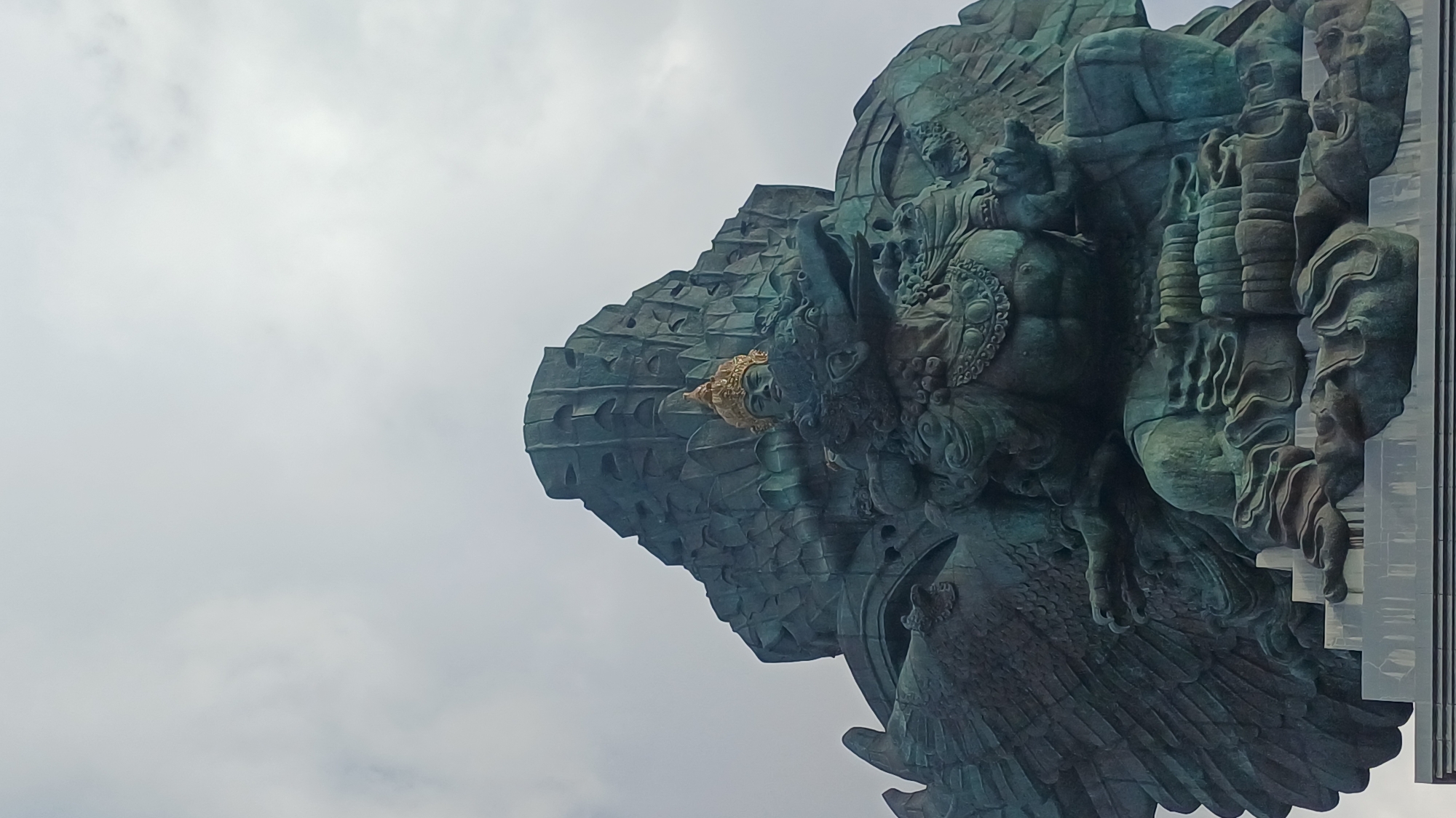 A low-angle shot captures a portion of the Garuda Wisnu Kencana statue against a backdrop of overcast sky in Kuta Selatan, Indonesia. The weathered, greenish-bronze statue features intricate carvings and textures, notably wings, faces, and ornate garments. A golden crown adorns one of the figures. The statue's design includes multiple layers and levels, with some sections appearing to be constructed from modular elements or panels.