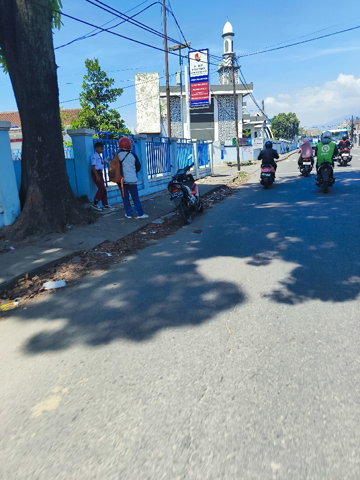 The image shows a street scene in Margahayu, Indonesia, at the coordinates -6.982 latitude and 107.563 longitude. The scene is in front of a building with a blue and white fence and a mosque behind it. The street is paved with asphalt. Three men on motorcycles are riding towards the camera, and two boys are standing near the fence. The boys are wearing school uniforms. The street is mostly empty, except for the motorcycles and the two boys. The sky is blue with a few clouds. The sun is shining, casting long shadows from the building and the people. A large tree stands on the left side of the image, creating a shadow across the sidewalk and street. There is debris on the sidewalk, including a paper bag and a discarded branch. The building behind the fence appears to be some type of business. A sign on the building has the text "A. RM. HARI - BAKSO" on it.
