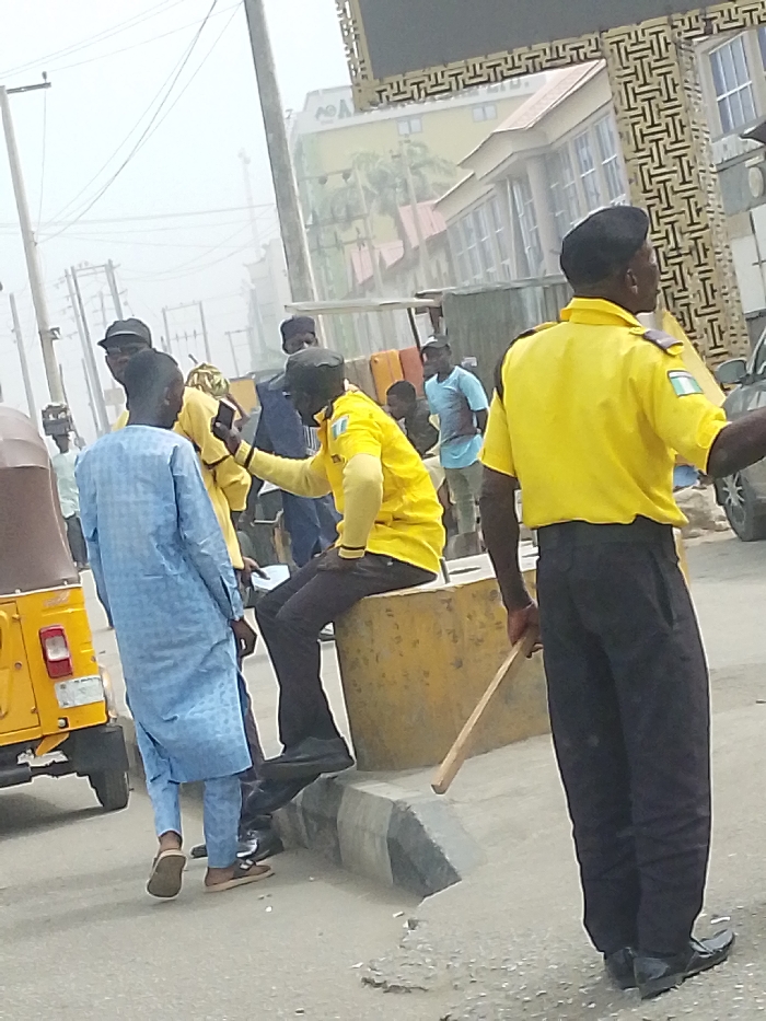 The image captures an outdoor urban street scene in Gandu Albasa, Nigeria, under a hazy sky. In the immediate foreground, a male individual wearing a yellow short-sleeved uniform shirt with a Nigerian flag patch on the left arm, dark trousers, and a black beret stands with his back largely to the viewer, holding a wooden stick in his right hand, extending downwards. To his left, another male, similarly dressed in a yellow uniform shirt, dark trousers, and a black beret, is seated on a large, rectangular yellow concrete barrier or curb block. This seated individual holds a smartphone in his right hand, oriented towards a male in a light blue patterned long tunic (agbada) who stands facing away from the camera. A yellow three-wheeled auto-rickshaw is partially visible on the far left. Multiple other male individuals in civilian attire are present in the mid-ground and background, some appearing to be standing or conversing. The background reveals various urban structures, including a multi-story building with a gold-colored facade and a visible "AO^" logo, and another building featuring a decorative geometric patterned exterior. Overhead utility poles and electrical lines crisscross the pale sky. The asphalt road surface exhibits minor wear, and a concrete curb line is visible.