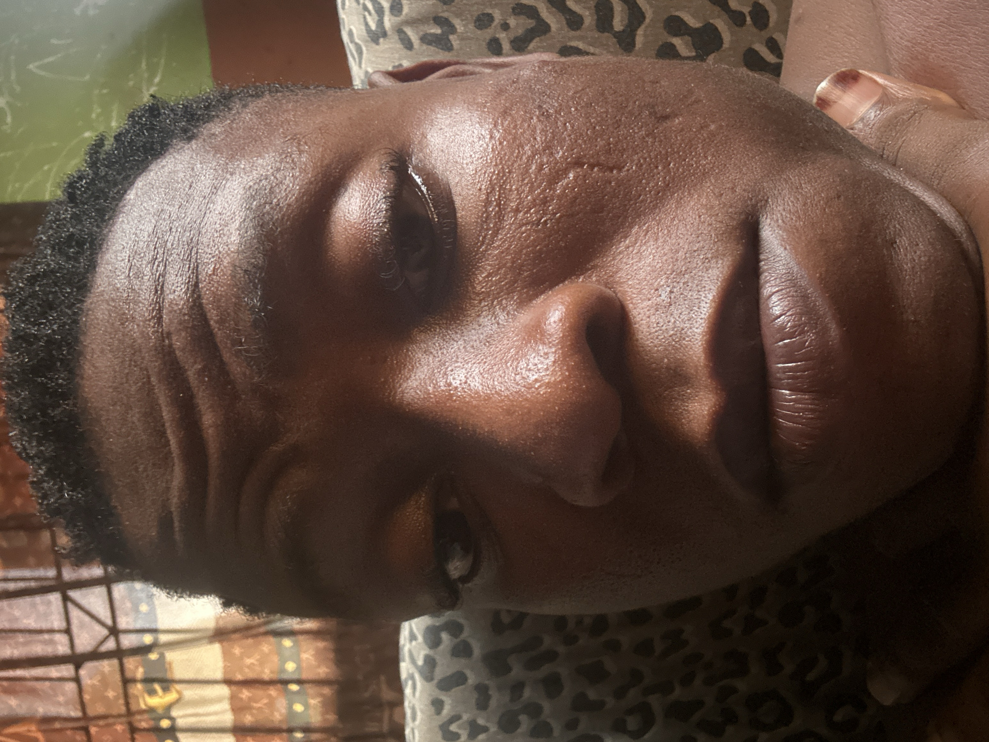 This image is a close-up selfie of a Black individual, seemingly taken indoors, as suggested by the presence of a leopard-print patterned cushion and a curtain with vertical stripes and horizontal yellow and brown bands in the background. The individual's face is the primary subject, with a focus on their features. Their skin appears moist, possibly due to sweat or tears, with prominent wrinkles on the forehead and a visible tear track on the cheekbone. The lighting highlights the texture of the skin and creates shadows that emphasize the contours of the face, including the nose and lips. The hair is short and dark, with a textured, curly appearance. The individual's eyes are partially closed, and their lips are slightly parted. The overall mood of the image could be interpreted as somber or introspective due to the facial expression and the glistening skin. The specific location within Ikorodu, Nigeria, cannot be determined from the visual information provided in the image itself.