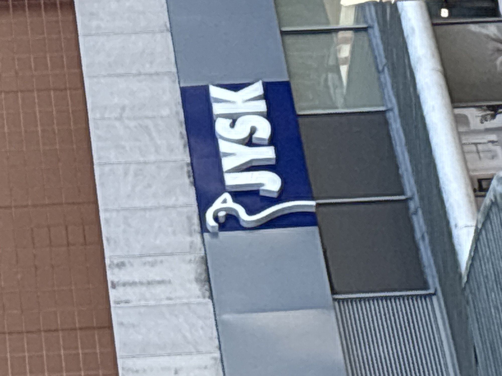 The image shows a sign for "JYSK" on the exterior of a building. The sign is illuminated with white lettering on a dark blue background. Above the JYSK sign, there is a light-colored textured wall, and above that, a brown brick wall with a grid pattern. Below the sign, there are windows and a gray section of the building. The scene is consistent with the JYSK store located at **24-25 St. Patrick's Street, Cork, Ireland**.