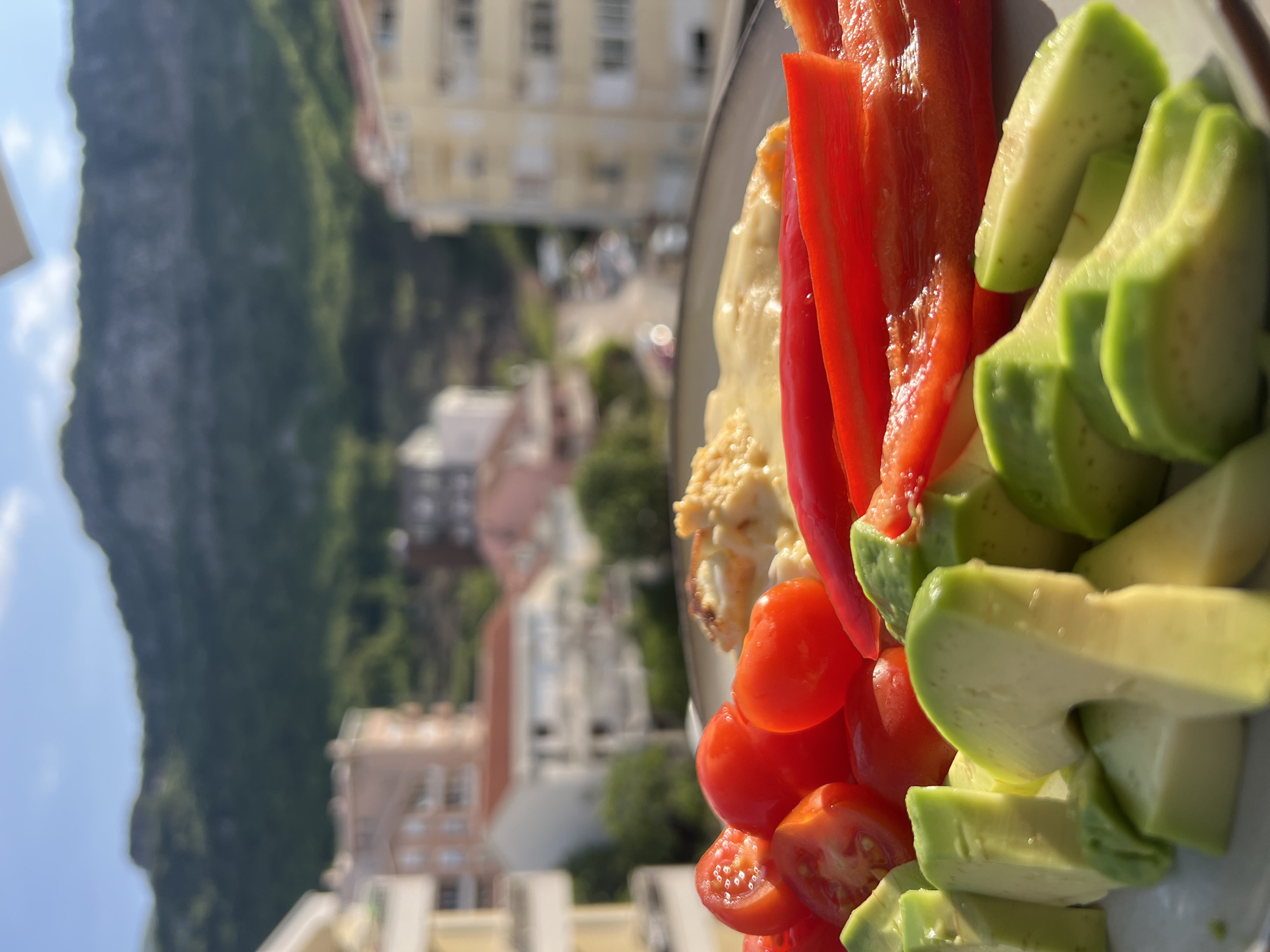 The image displays a vibrant meal placed outdoors, likely on a balcony or terrace, with a scenic view in the background. The primary subject is a plate filled with colorful, healthy food. On the plate, there are bright red slices of bell pepper, neatly cut green avocado slices, and small, ripe red tomatoes, some whole and some halved. Adjacent to these vegetables is a creamy, textured dish, possibly scrambled eggs or a similar preparation, with a slightly golden hue.
The background reveals a picturesque outdoor setting identified as Budva, Montenegro. To the left, a large, dark green, tree-covered mountain or cliff face dominates the landscape under a clear, bright sky with scattered clouds. Below and to the right of the mountain, various multi-story buildings of different architectural styles are visible, suggesting an urban or semi-urban environment. The sunlight is strong, indicating a clear day, likely morning or late afternoon, as it casts distinct shadows and highlights the food on the plate. No people or visible text are present in the image. The scene captures a moment of an inviting meal with a stunning natural and architectural backdrop.