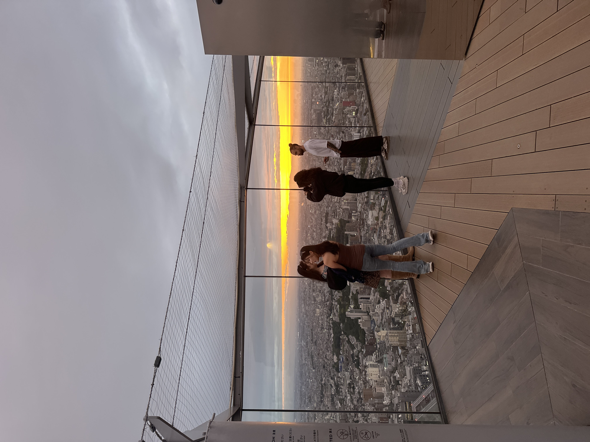The image shows four people on an outdoor platform, presumably a high-rise observation deck, overlooking a cityscape at sunset. The location is Shibuya, Japan, recognizable by the urban layout and architecture. The platform has a wooden deck and a glass barrier with a protective net. The sunset paints the sky with hues of orange and yellow, visible through the glass and illuminating the cityscape below. The first person is in a white shirt and black pants. The second person is wearing a black jacket. The third and fourth people are standing closer, one in a brown top and jeans. They are facing outward, presumably looking at the sunset or the cityscape.
