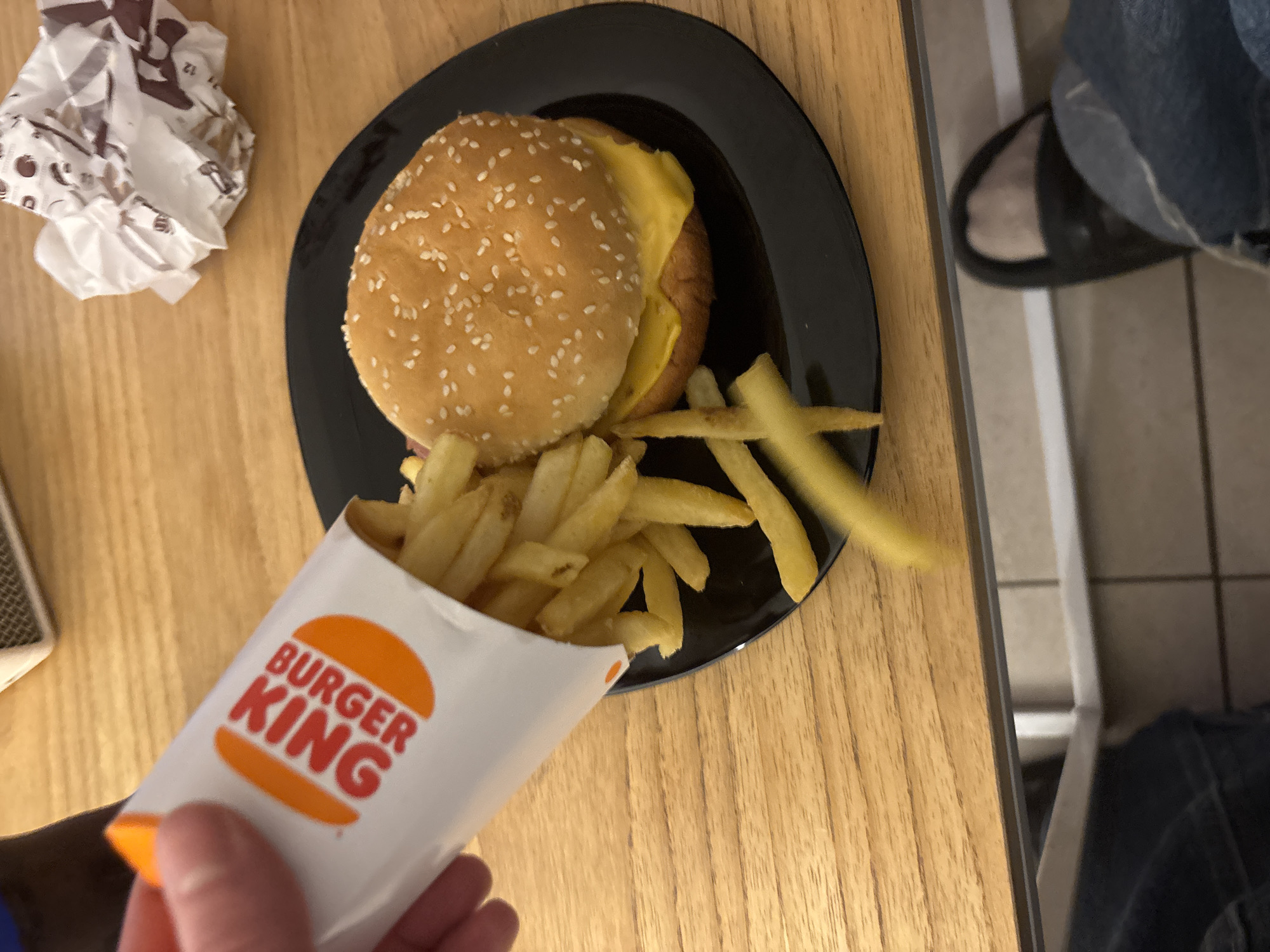 A partially consumed Burger King meal is displayed on a light brown wooden table. The meal consists of a burger, predominantly visible with its sesame seed bun and a visible slice of yellow cheese, resting on a black plate. A serving of fries, partially contained within a Burger King branded paper container, spills onto the plate. To the left of the plate is crumpled Burger King wrapper paper. The scene is interrupted at the right edge by the presence of a person's leg, clad in denim jeans, with a black and grey slipper on their foot. The location is unidentified, and there is no visible evidence to link the image to the city of Buenos Aires, Argentina.