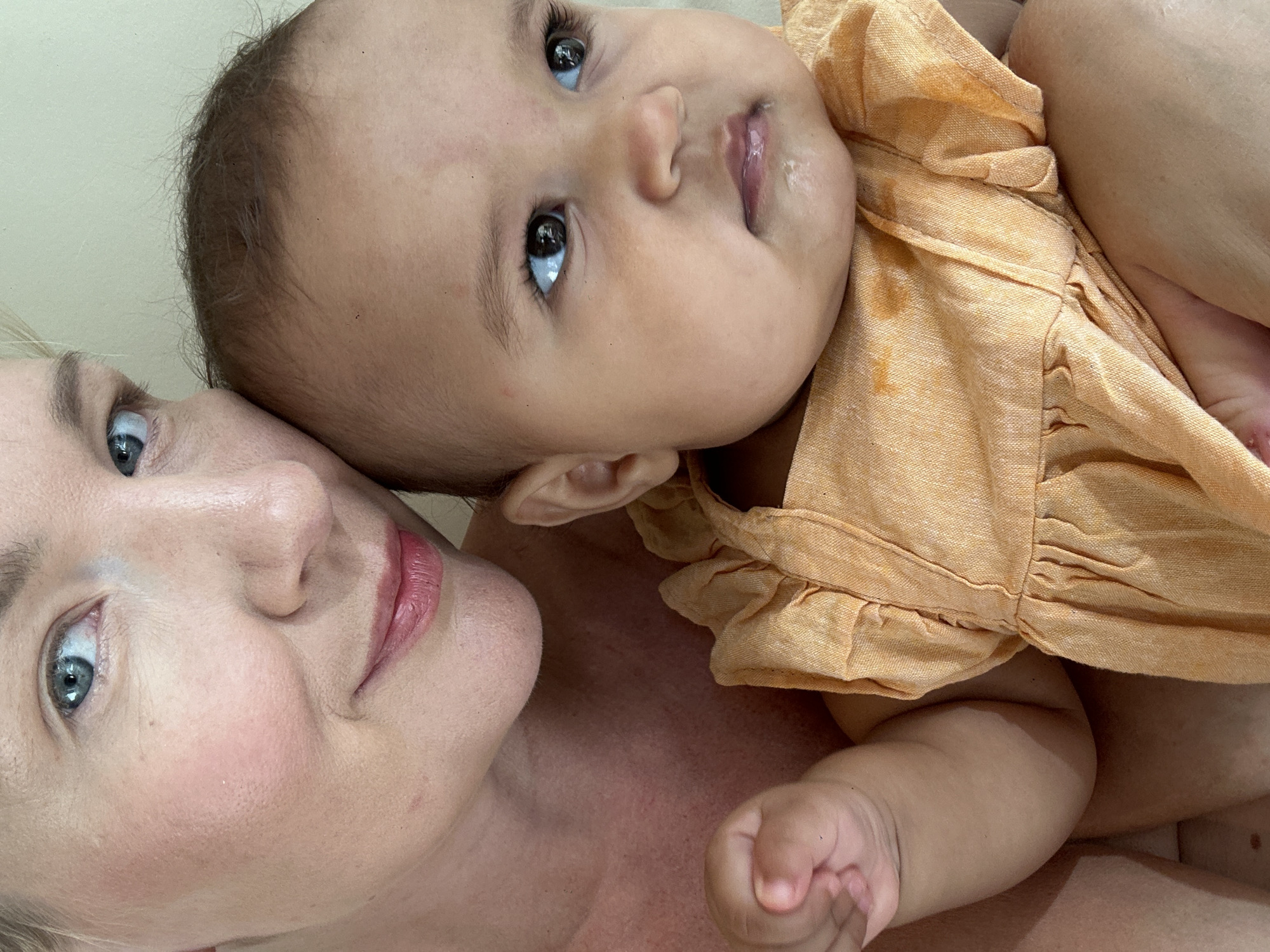 A close-up shot shows a Caucasian woman lying next to and holding a baby. The woman has fair skin, blue eyes, and pink lips. The baby has brown hair, dark eyes, and appears to be wearing an orange dress. The background is a plain light-colored surface. There is no indication of the location being North Kuta, Indonesia, or any specific city.