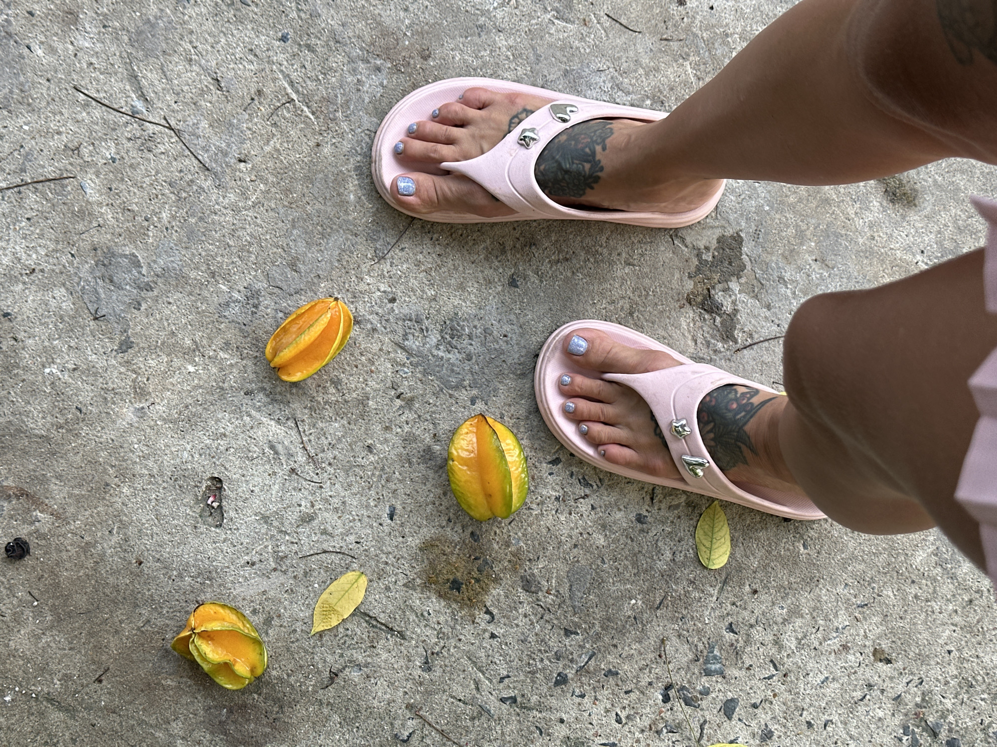 The image depicts a high-angle view of a person's lower legs and feet on a concrete surface in Mũi Né, Vietnam. The individual wears light pink flip-flops. Both feet feature light blue or silver glitter nail polish on the toenails. Dark, intricate tattoos are visible on the top of each foot, extending towards the ankles. Three star fruits (carambola), varying from yellow-green to orange, are scattered on the rough, light gray concrete ground. Several dried, yellow-brown leaves and small debris are also present on the surface. The scene is illuminated by natural light, casting subtle shadows.