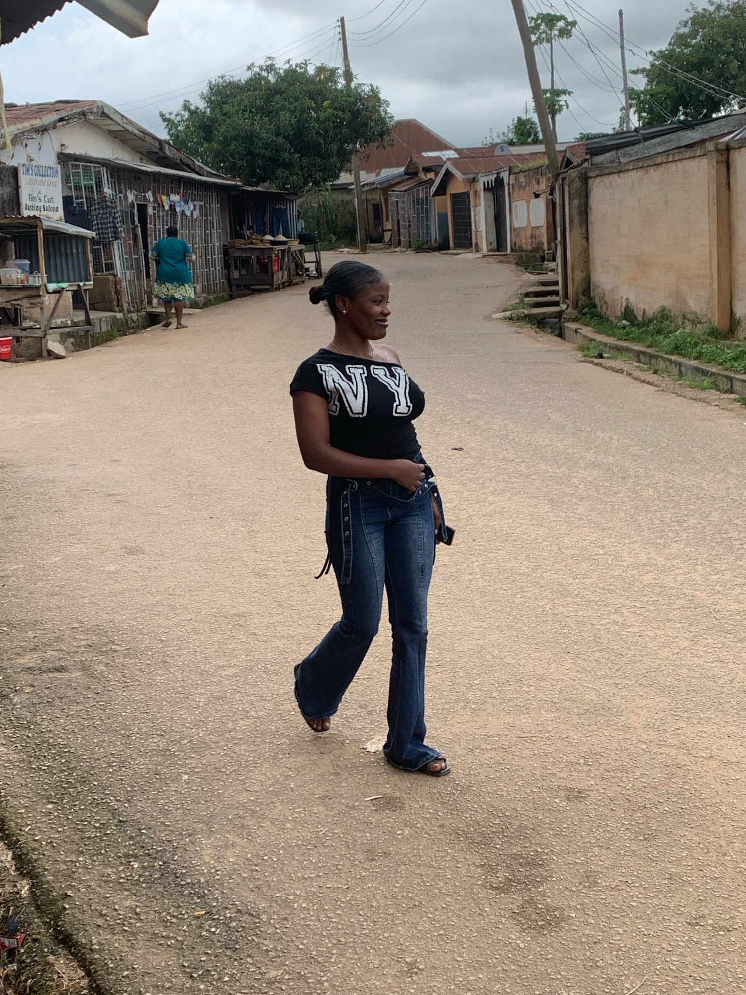 In Jos, Nigeria, a young woman is captured mid-stride on a paved road, her gaze directed forward. She is dressed in a black t-shirt with a white "NY" logo and blue bell-bottom jeans, accessorized with dark sandals. Her dark hair is pulled back in a bun. The setting is a street lined with modest buildings, some appearing to be shops or homes, with corrugated metal roofs visible. A woman in a green dress walks away from the camera in the background, near a shop displaying clothes and other items. The road surface is textured asphalt, with some patchy discoloration and scattered debris. The environment appears to be a developing urban area, with overhead electrical wires present and a cloudy sky overhead.
