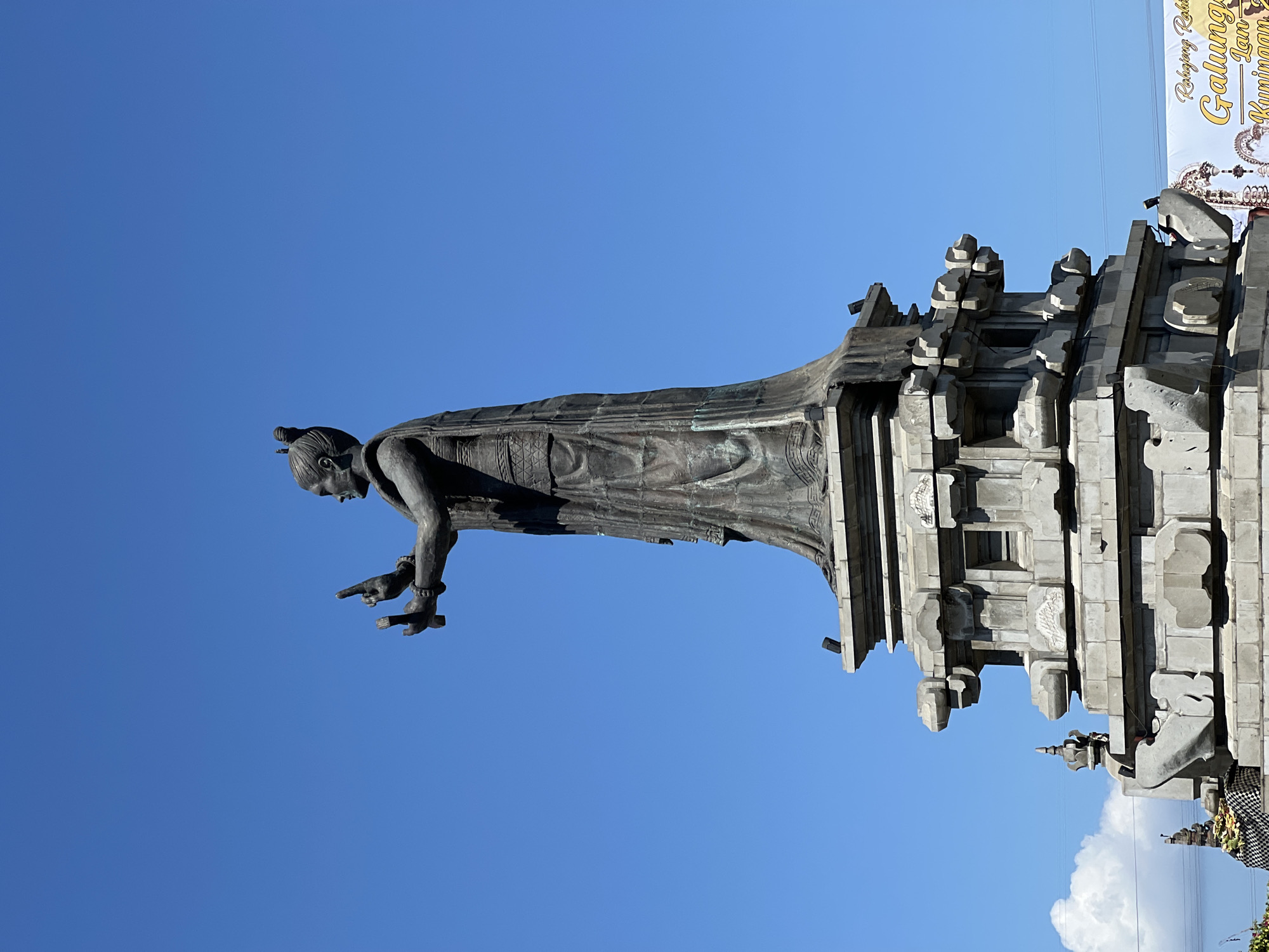A full shot captures a bronze statue of a woman in traditional attire, with her hands outstretched and holding what appear to be small objects. She stands atop a multi-tiered stone pedestal adorned with intricate carvings. The statue and pedestal are set against a clear blue sky. In the lower right corner of the image, a white banner with yellow text and decorative elements is partially visible. Below the pedestal, a portion of a stone structure with decorative elements and what looks like offerings can be seen on the left. The overall scene suggests a cultural or religious monument in an outdoor setting, likely in the city of Dawan, Indonesia, given the hint.