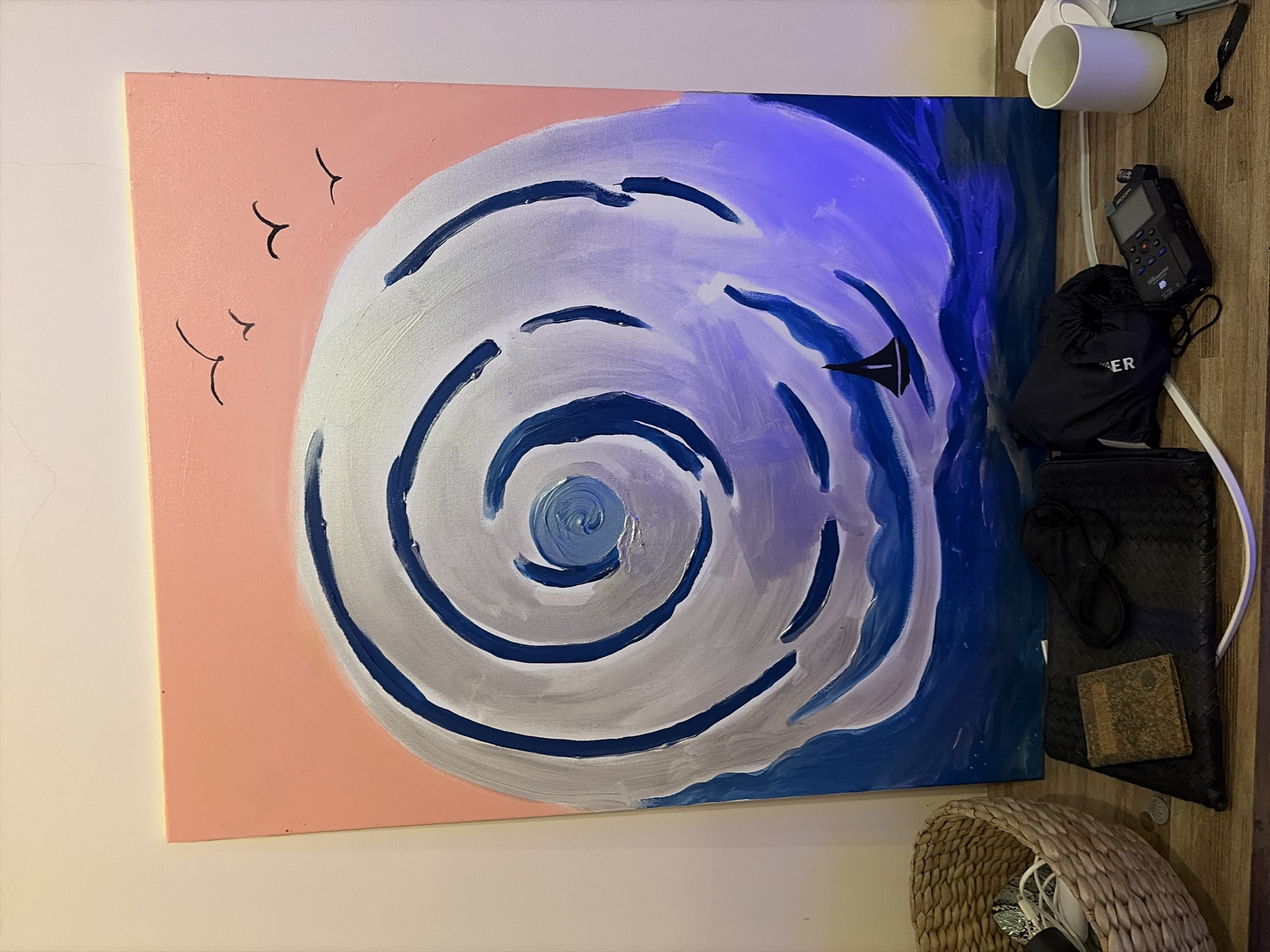 The image shows a painting of a swirling wave with a sailboat in the foreground. The painting is on an easel and leans against a wall. There are several objects on a wooden surface in front of the painting, including a woven basket, a wallet, a black pouch, a white mug, and a black digital recorder. The lighting in the scene is a mix of natural and artificial light, with a blueish hue cast on the right side of the painting. The scene appears to be indoors, on a shelf or table. There is no discernible location in the city of Mengwi, Indonesia.