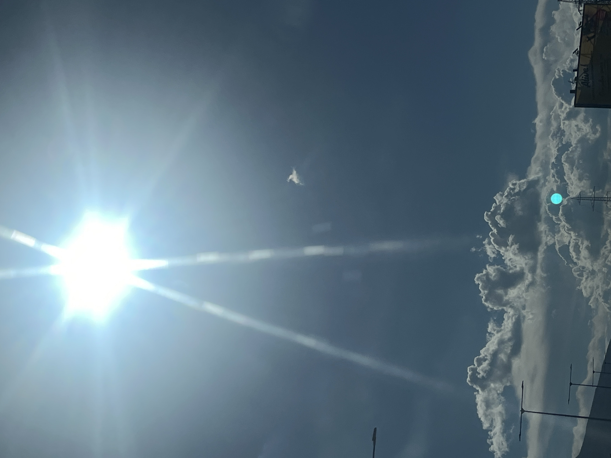 A bright sun shines in a clear blue sky. There are some clouds in the lower part of the image. A faint object is visible in the sky, possibly a bird or a piece of debris. A billboard is visible on the right side of the image. There are also some streetlights in the lower left part of the image. The image is taken from a low angle, looking up at the sky.