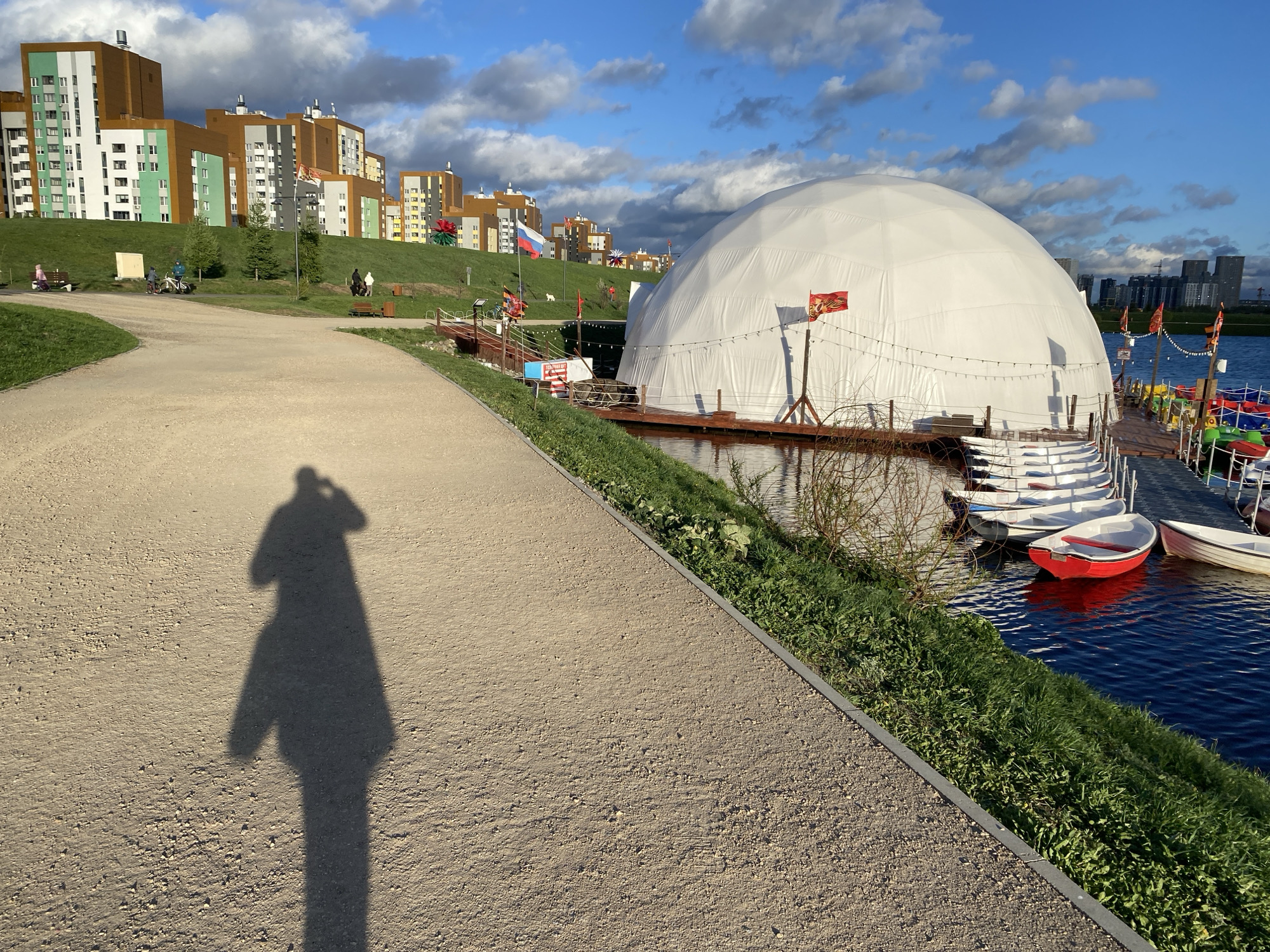The image shows a park in Yekaterinburg, Russia, on a sunny day with scattered clouds. A wide, gravel path runs along the lower left of the frame, casting a long shadow of a person holding a camera. To the right of the path is a strip of grass bordering a body of water. A large, white dome structure is situated on a pier extending into the water. Several small boats, some red and white, are docked next to the pier. In the background are colorful, modern apartment buildings on a grassy hillside. Flags are visible on the pier and near the buildings. People can be seen walking and cycling in the background.