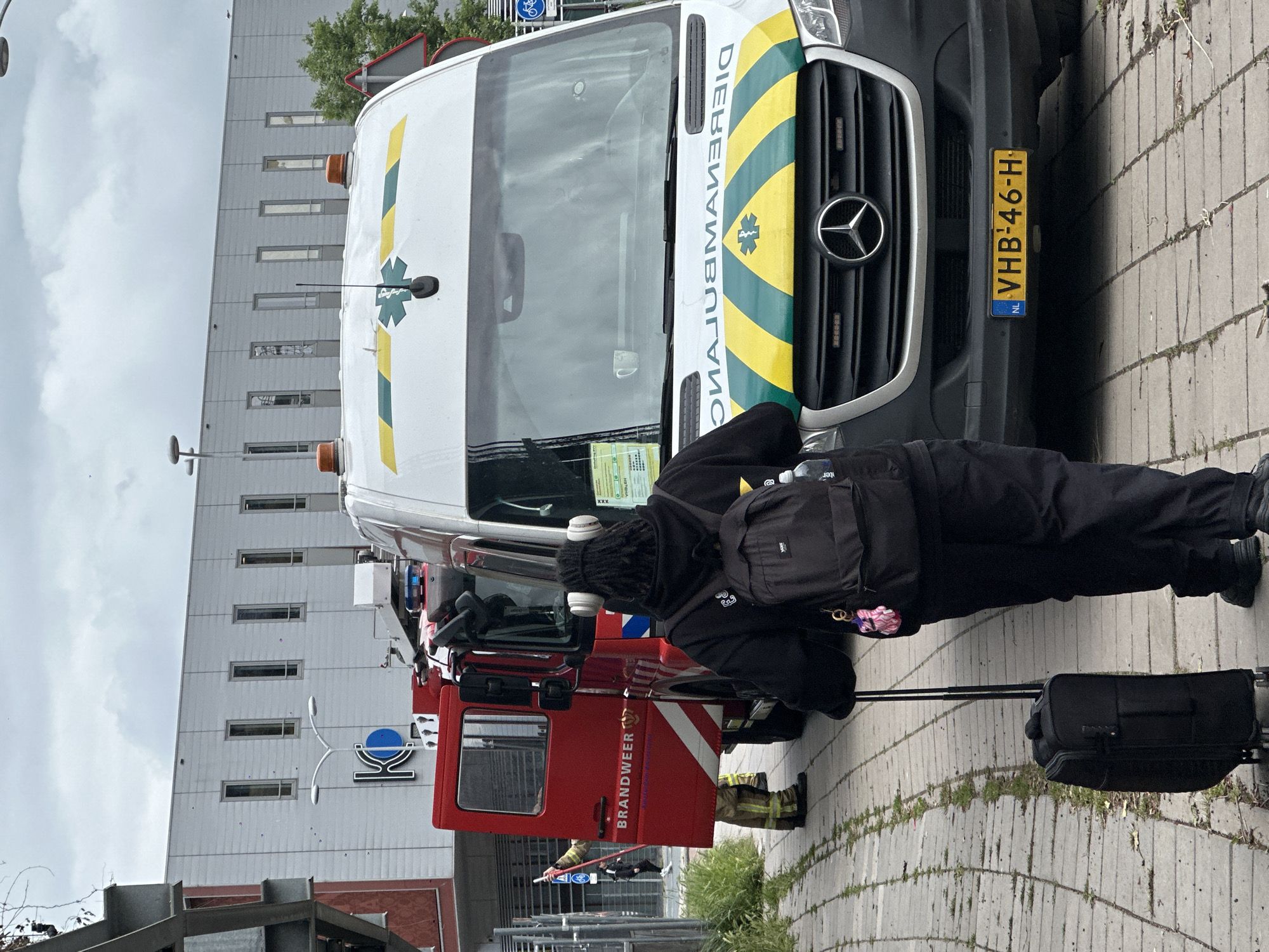 An image shows a person in black clothing, wearing headphones and carrying a black backpack and a black rolling suitcase, standing on a paved walkway in front of an ambulance and a red fire truck. The fire truck has "BRANDWEER" written on its side. The ambulance has a green and yellow stripe design and a star of life emblem. A person in a firefighter's uniform is visible near the open door of the fire truck. A building with a modern facade is in the background. A street sign with a bicycle symbol is visible to the right of the ambulance. The license plate of the ambulance reads "VHB-46-H". The scene appears to be a street in Amsterdam, Netherlands.
