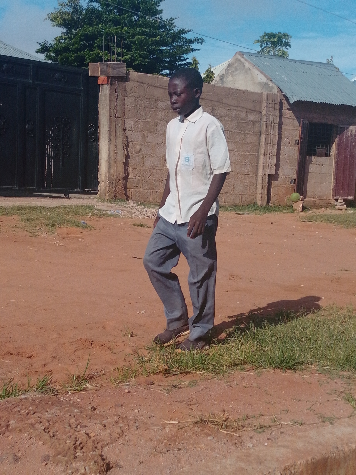 A young African boy, appearing to be around 10-12 years old, walks on a dirt path in Gombe, Nigeria. He is wearing a light-colored collared shirt with a logo on the left breast and gray trousers. The boy is walking from left to right in the frame, with his head turned slightly to his left, looking downwards. He is wearing dark shoes. The background consists of a brick wall, a black metal gate, and a corrugated metal roof structure. There is a green ball visible near the wall. The ground is predominantly reddish-brown dirt with patches of dry grass. The sky is blue with some clouds. The lighting suggests it is daytime.