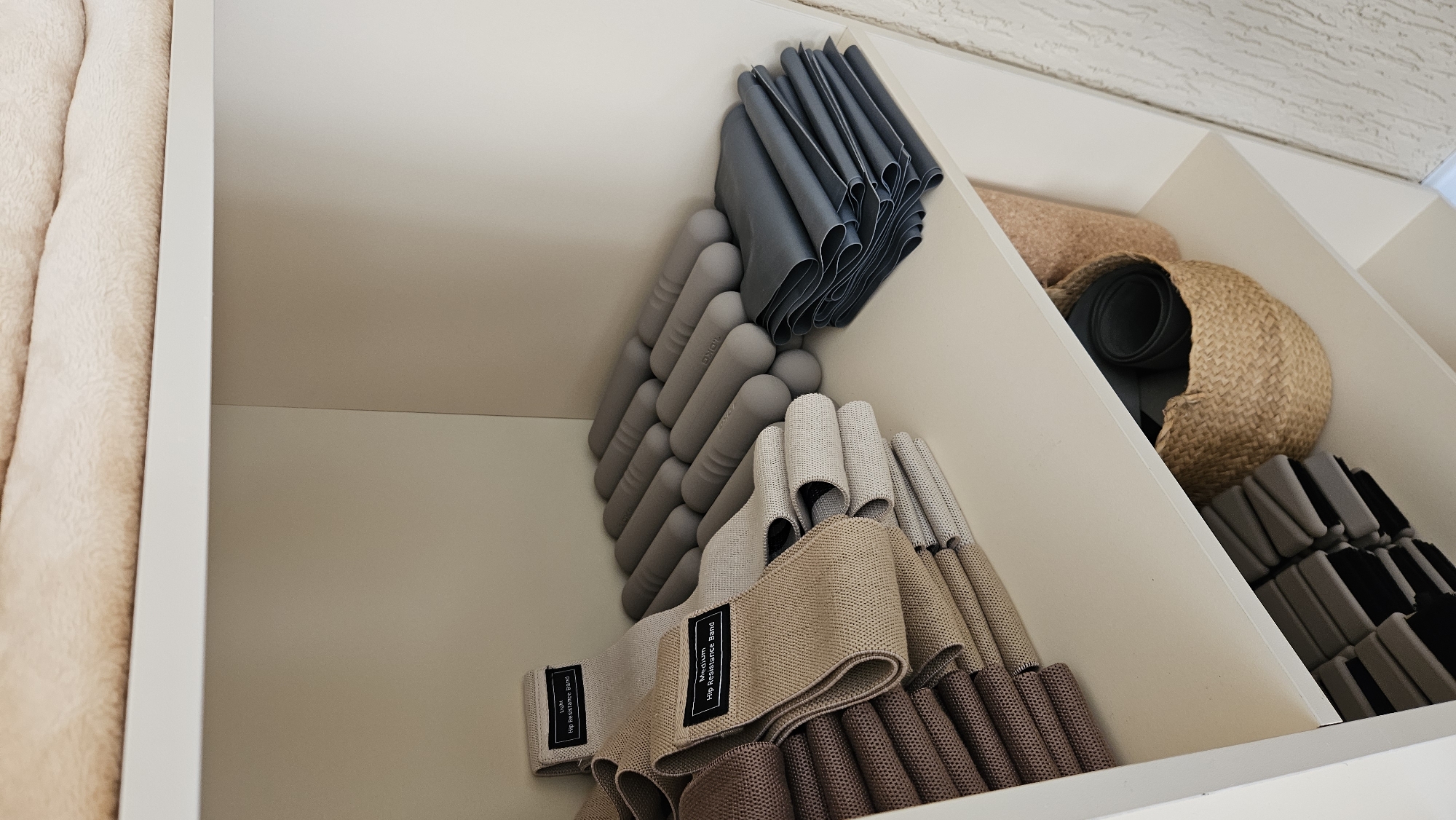 The image shows an organized shelf unit filled with fitness equipment. On the top shelf, there are folded beige and brown fabric resistance bands, with labels indicating "Light Hip Resistance Band" and "Medium Hip Resistance Band". Next to the bands, there are rows of gray cylindrical foam rollers. On the right side of the top shelf, a stack of dark blue folded fabric resistance bands is visible. The middle shelf contains a woven basket filled with rolled-up dark gray resistance bands, and a taller, light brown woven container. The bottom shelf is partially visible and appears to hold more stacked resistance bands. The background is a plain wall. The scene is indoors, within a storage space. There is no visible indication of the city of Vladimir, Russia.