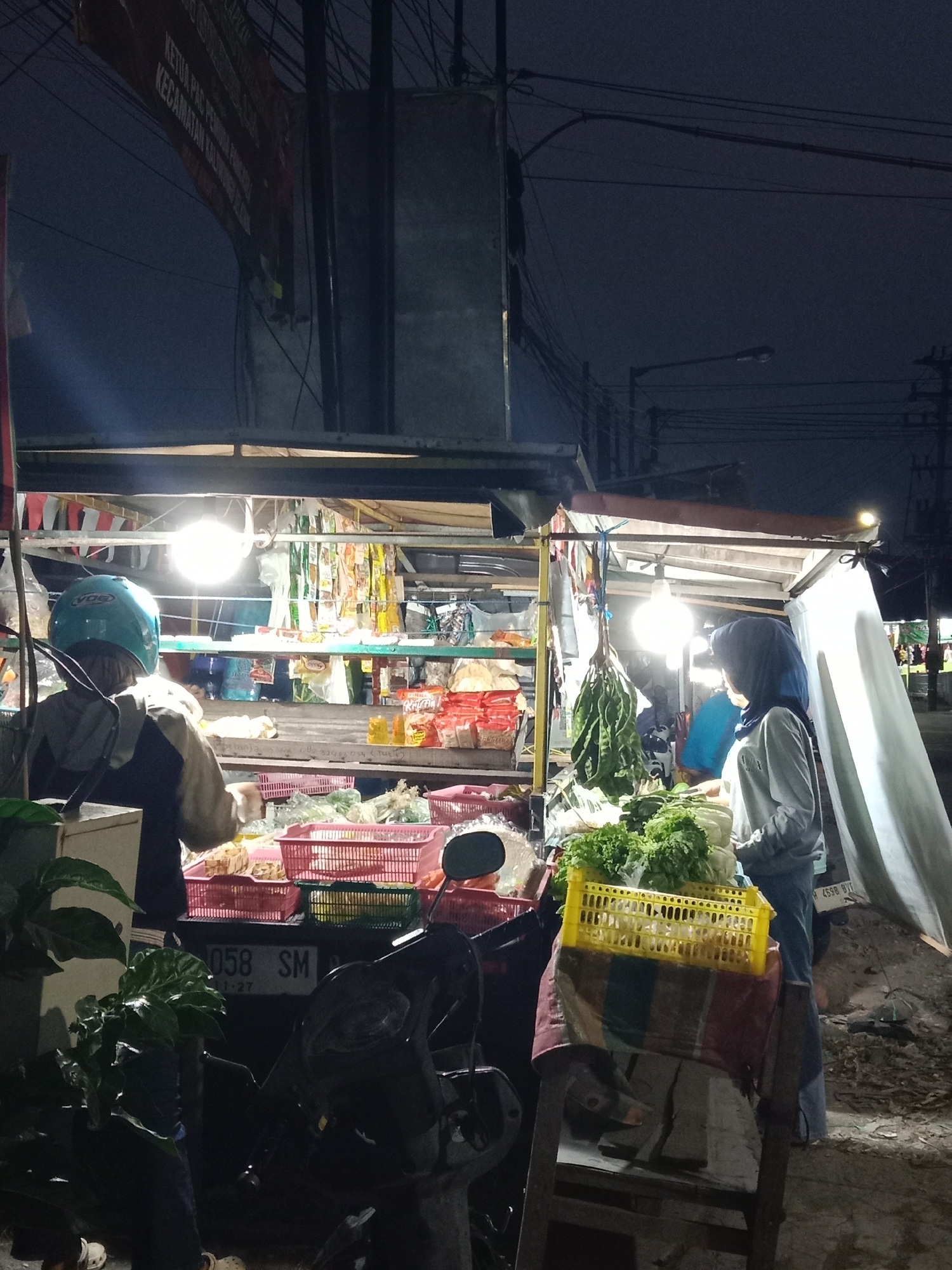In the city of Langensari, Indonesia, at night, a street vendor stall displays various goods. Two individuals, likely a vendor and a customer, are present. The stall is illuminated by bright lights, highlighting rows of packaged items, fresh produce like lettuce and possibly bitter beans, and baskets of what appear to be smaller vegetables or fruits. A motorcycle is parked in front of the stall, its license plate partially visible. Overhead, power lines and streetlights are visible against the dark sky. One person wears a blue helmet and a grey jacket, while the other is dressed in a blue hijab and a grey top. The scene suggests a local market operating in the evening.