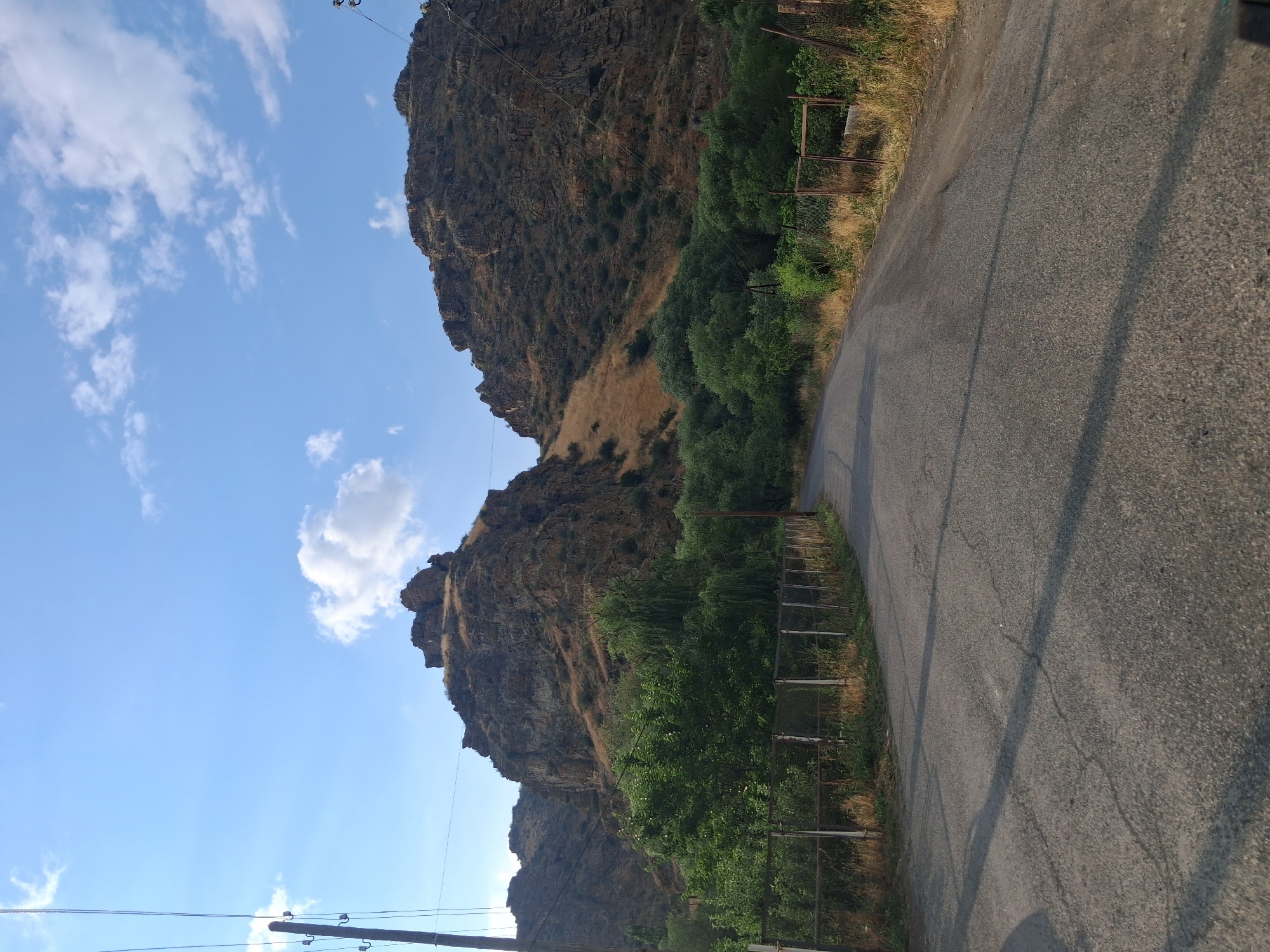 The image shows a paved road winding through a rural landscape with a dramatic rocky mountainside dominating the background. The mountains are steep and rugged, covered in dry grass and sparse vegetation. A line of trees, predominantly green and leafy, runs along the base of the mountains, separating them from the road. A wire fence runs along the side of the road, bordered by dry grass and some scattered bushes. Power lines and poles are visible on the left side of the image, stretching upwards towards the blue sky, which is dotted with a few white clouds. The road appears to be in moderate condition, with some visible cracks and patches. The overall scene suggests a remote, natural environment. The location is likely in or near the city of Vayk, Armenia, given its mountainous terrain and rural character.