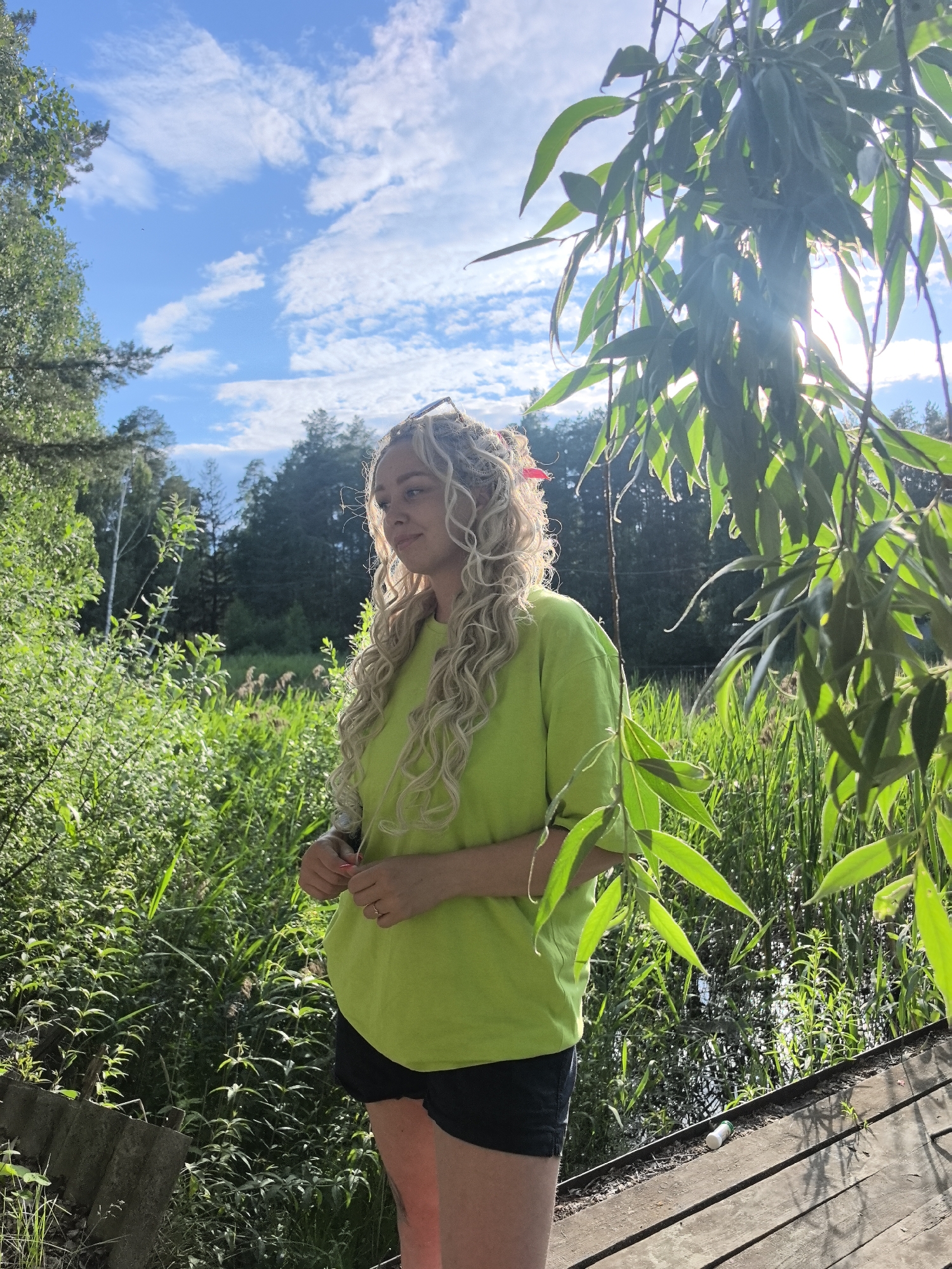 The image depicts a young woman with blonde, curly hair standing outdoors in a natural environment. She is wearing a bright lime green t-shirt and black shorts. Her body is positioned facing right, with her head turned to the left, looking into the distance. She has a slight smile on her face. The background consists of lush green foliage, reeds, and trees, suggesting a rural or park-like setting. The sky above is blue with scattered white clouds. Sunlight filters through the leaves of a tree in the foreground, creating a dappled lighting effect. A wooden structure, possibly a dock or a walkway, is visible at the bottom right of the image. The scene appears to be set in the city of Noviy, Russia, as specified.