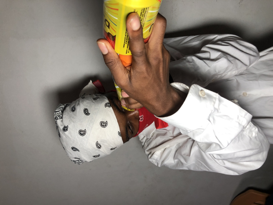 In Ngleshi Amanfro, Ghana, a single individual is depicted from the torso up against a plain grey wall. The person, with dark skin, wears a white long-sleeved shirt and a red bow tie. A white bandana with black paisley patterns covers the head, with a portion of red fabric visible beneath it on the left side. A red bandana covers the lower half of the face. The individual's dark-skinned hands hold a bright yellow cylindrical object, resembling a can or bottle with orange and red graphics and white text, horizontally to their face, positioned near the eyes and nose.