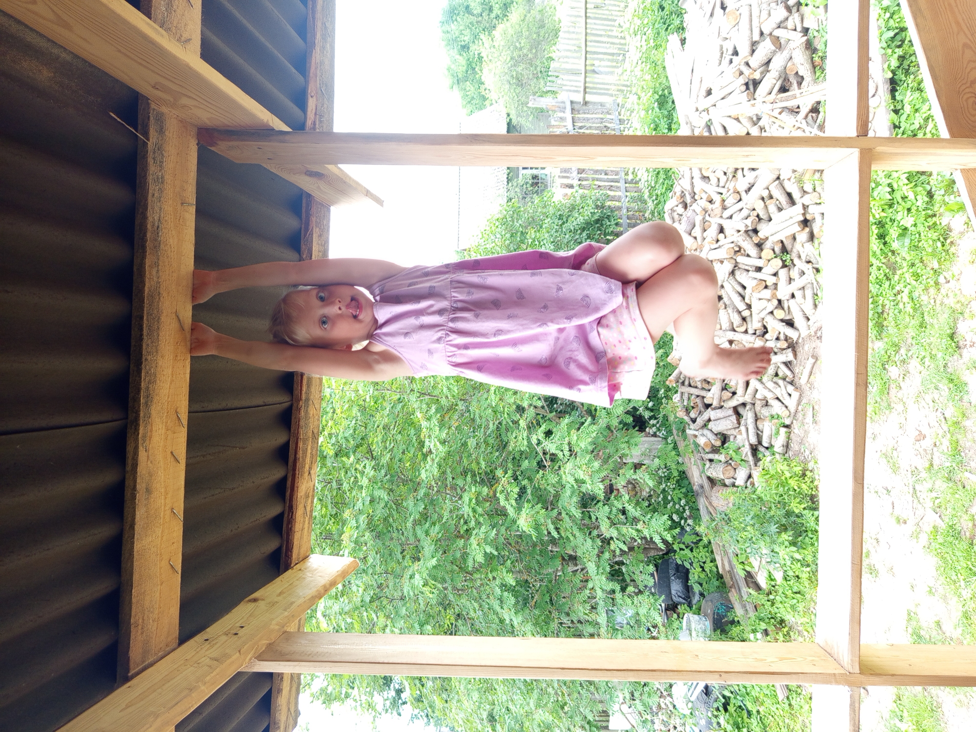 A young girl with blonde hair is hanging from a wooden beam with her hands. She is wearing a pink dress and patterned shorts. Her tongue is sticking out. The girl is in a semi-built wooden structure with a corrugated roof. In the background, there is dense green foliage, a pile of cut logs, and a wooden fence. The scene appears to be in a rural or suburban setting in Ostrovtsy, Russia, possibly a backyard or a construction site.