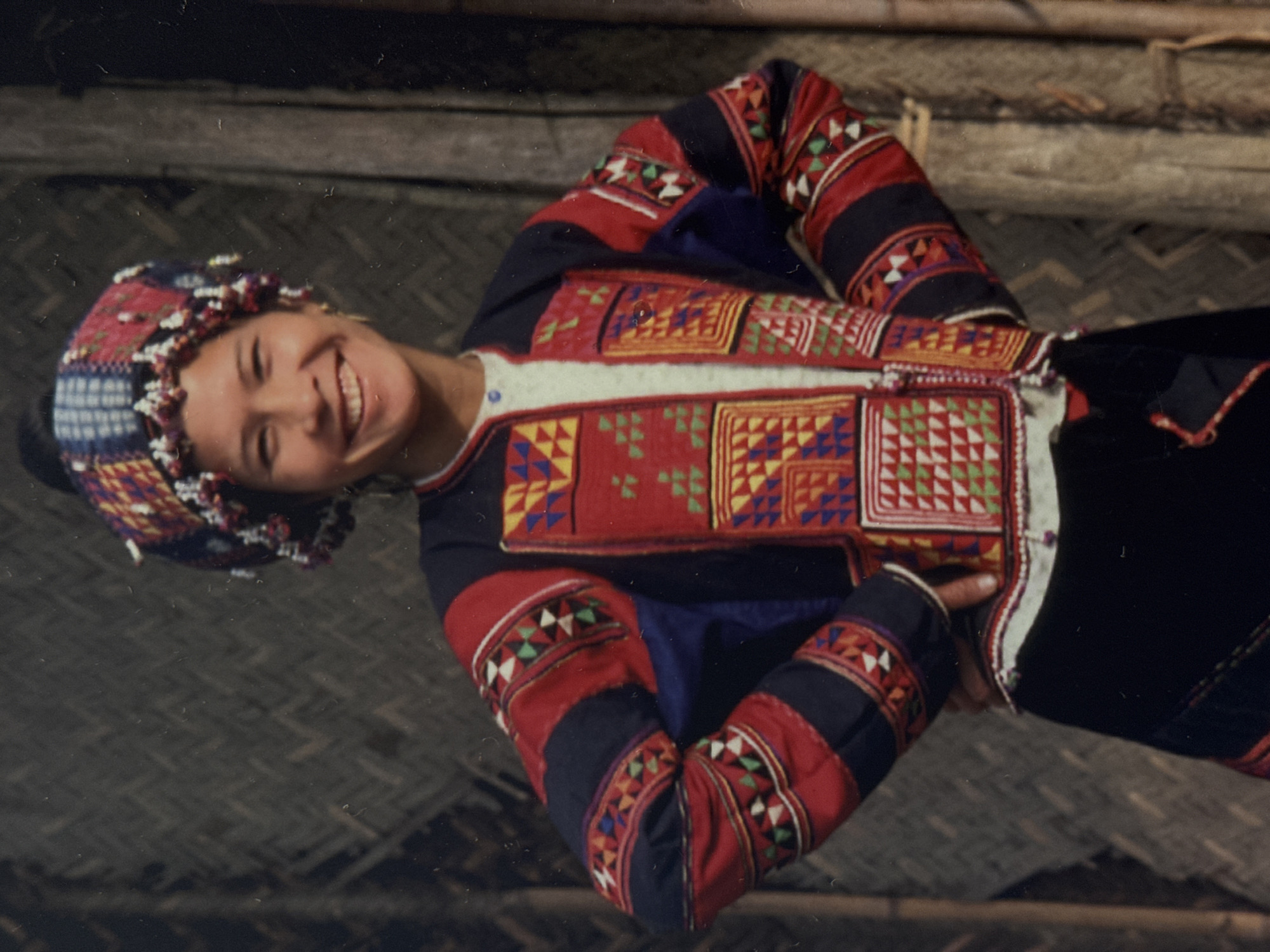 This is a vertically oriented, slightly blurry, color photograph of a young woman dressed in traditional clothing, likely from an ethnic minority group in Vietnam, given the location context of Hanoi. The woman is smiling broadly and looking towards the camera, her head tilted slightly. She is wearing an intricately embroidered jacket and a matching headdress. The jacket is predominantly red with black and navy blue accents, adorned with colorful geometric patterns in yellow, blue, green, and white, stitched in a patchwork style. The sleeves are also decorated with similar patterns. The headdress is a colorful band with beaded accents, possibly made of woven fabric. She is leaning on her arms, with her hands clasped in front of her chest. The background appears to be an interior setting, with wooden beams and woven mats, suggesting a rustic or traditional dwelling. The lighting appears to be natural, possibly daylight, but the exact time of day is not discernible. There is no visible text in the image. The overall mood is joyful and vibrant, highlighting the beauty of the traditional attire and the woman's cheerful expression.