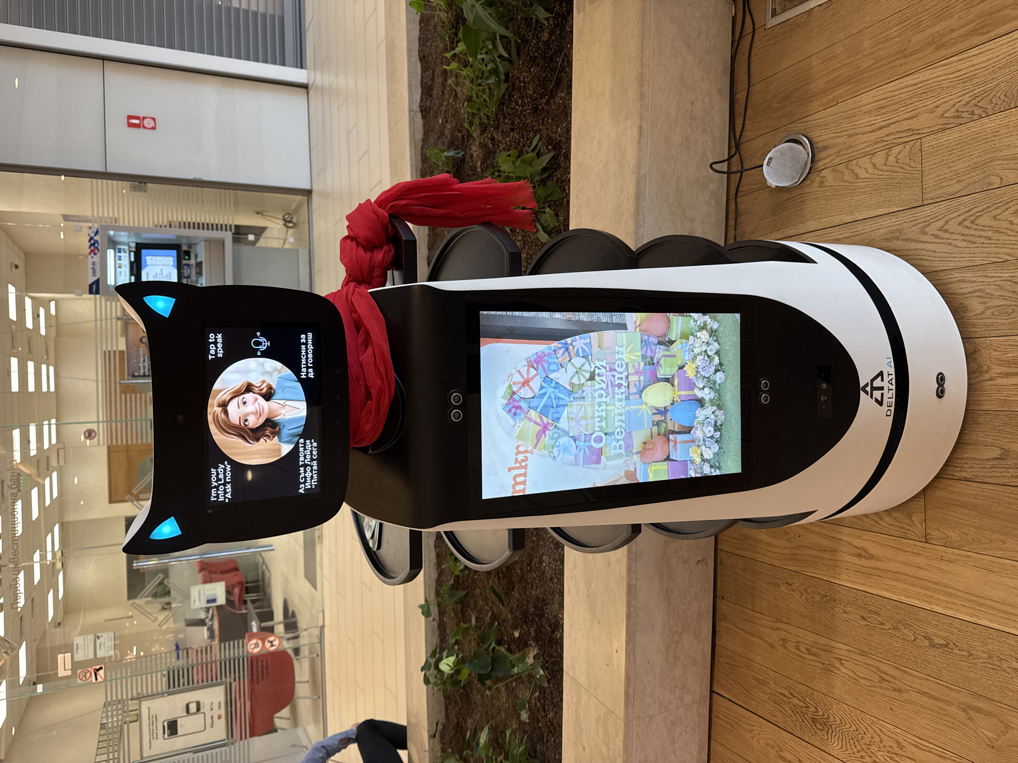 A close-up shot of a Deltatai informational robot located indoors, possibly in a shopping mall in Sofia, Bulgaria, is captured from a high angle. The robot has a white and black body, is equipped with two large screens showing promotional and information content, and features cat-like ear lights. A red scarf is draped around the robot's neck. The surrounding environment includes a wooden floor, an interior of a bank, and greenery. The robot is situated next to a wall. The first screen displays a cartoon-style woman with a microphone icon and a button that reads "Tap to speak." The second screen displays the text "mkp otkrii Velikden." The logo "DELTAT AI" is on the lower right-hand side of the robot's body.